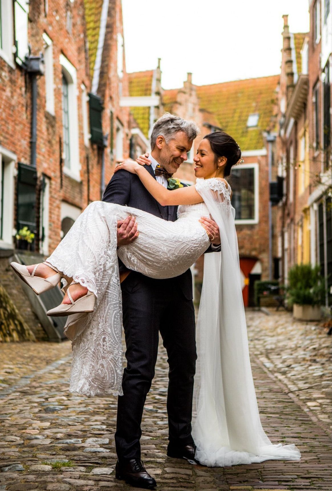 Bruidegom draagt ​​bruid in een geplaveid steegje. Ze glimlachen naar elkaar. De bruid draagt ​​een witte jurk.