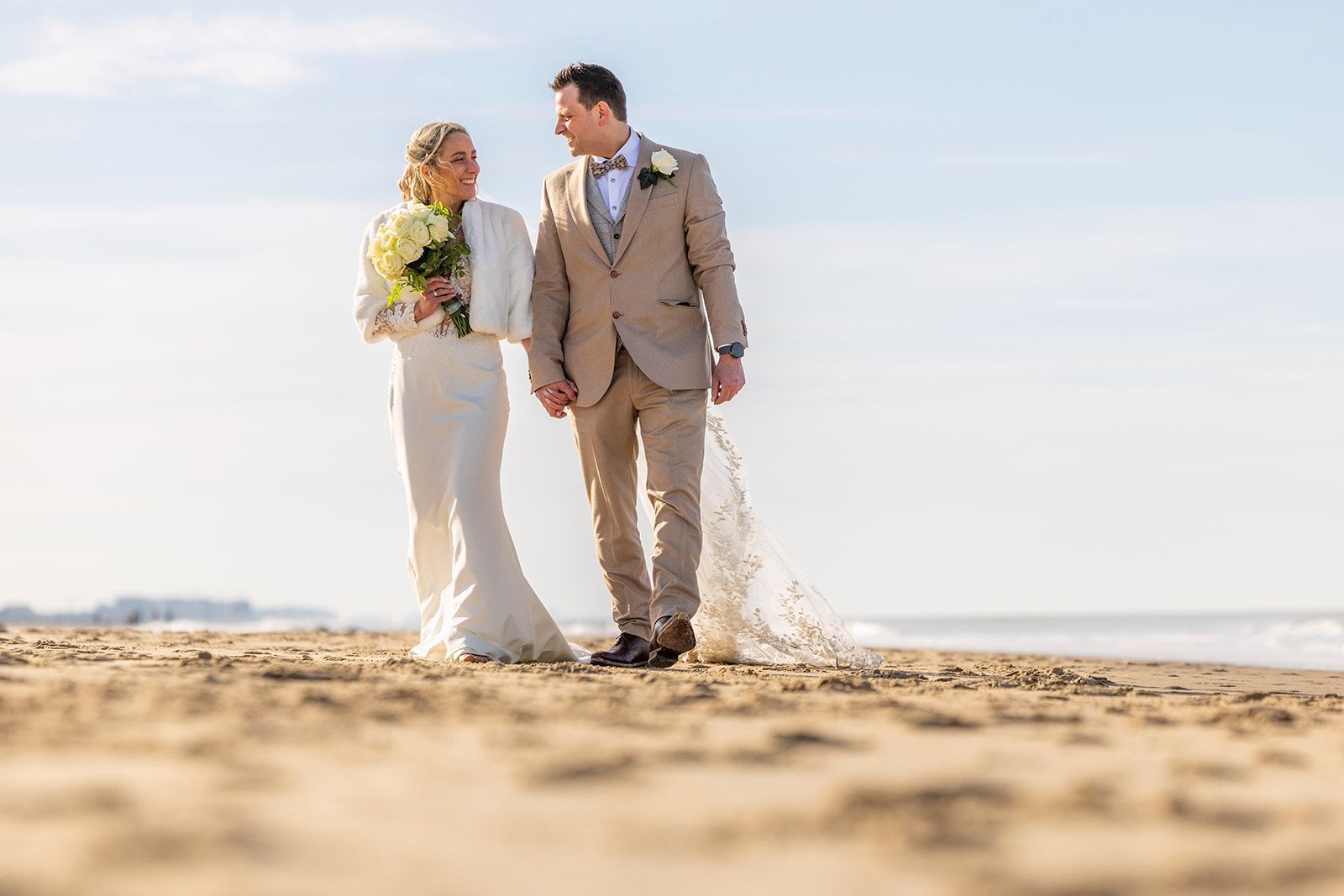 Een stel loopt hand in hand, glimlachend, over een pad; de bruid in een witte jurk, de bruidegom in een roestbruin pak.