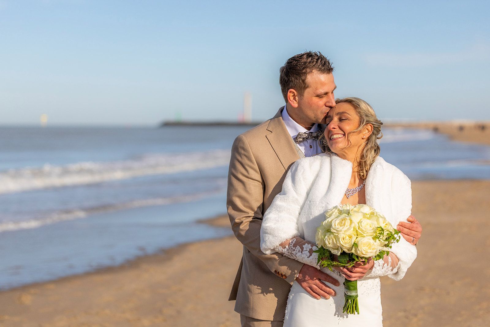 Pasgetrouwden omhelzen elkaar naast een klassieke zwarte auto. De bruid houdt bloemen vast, de bruidegom draagt ​​een rood pak; zonlicht in een voorstedelijke omgeving.