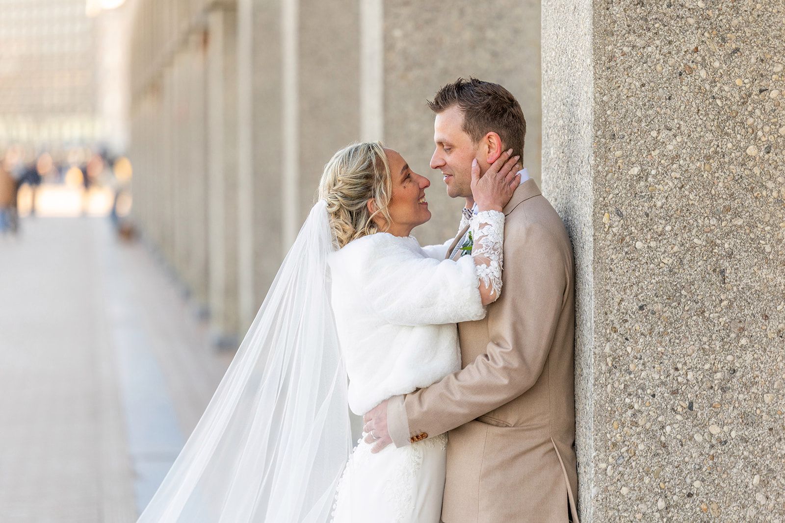 Een bruidspaar raakt elkaars voorhoofd aan en lacht voor een stenen gebouw.