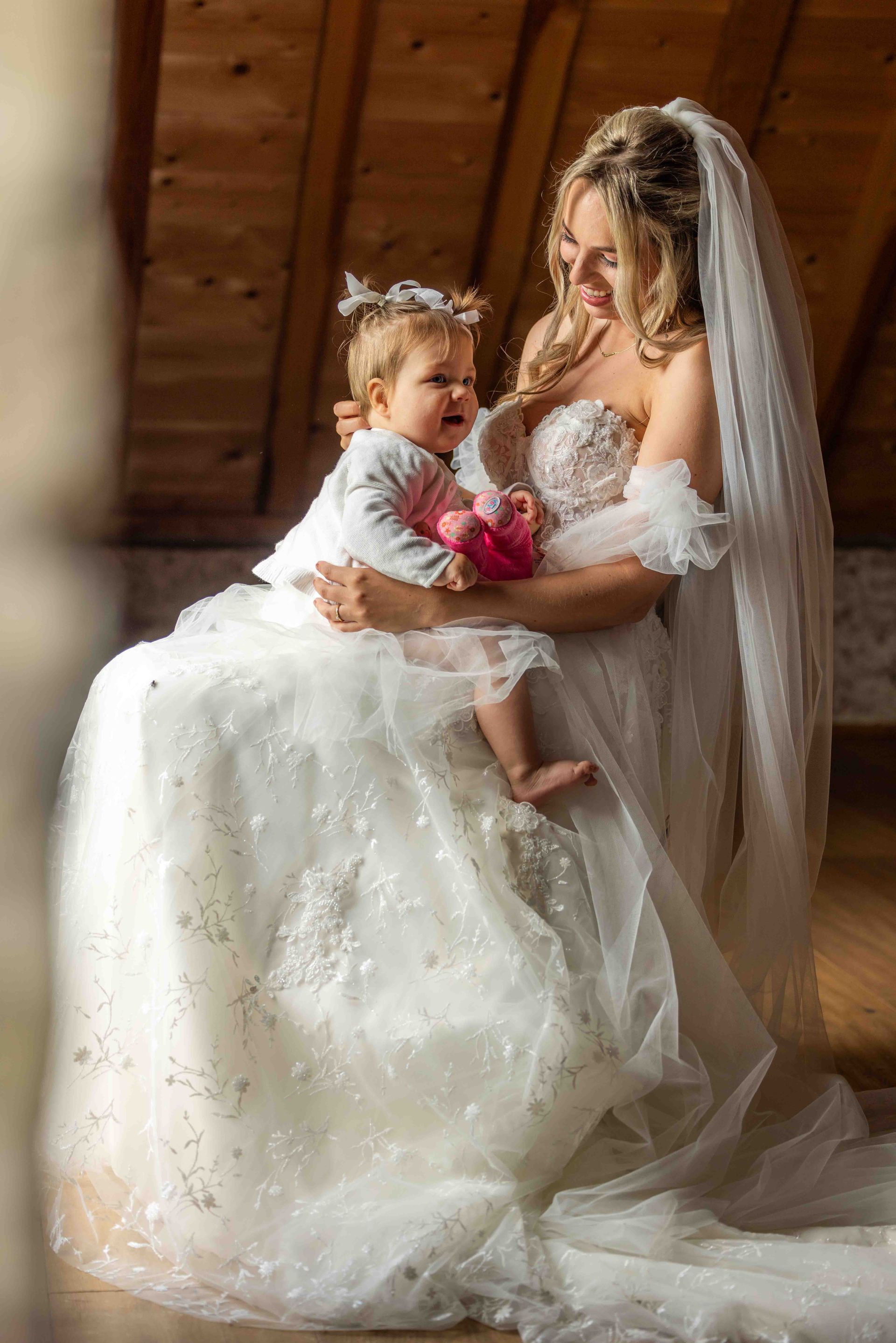 Een bruid in een witte jurk houdt een klein kind vast. Ze lachen allebei, in een gebouw met houten balken.