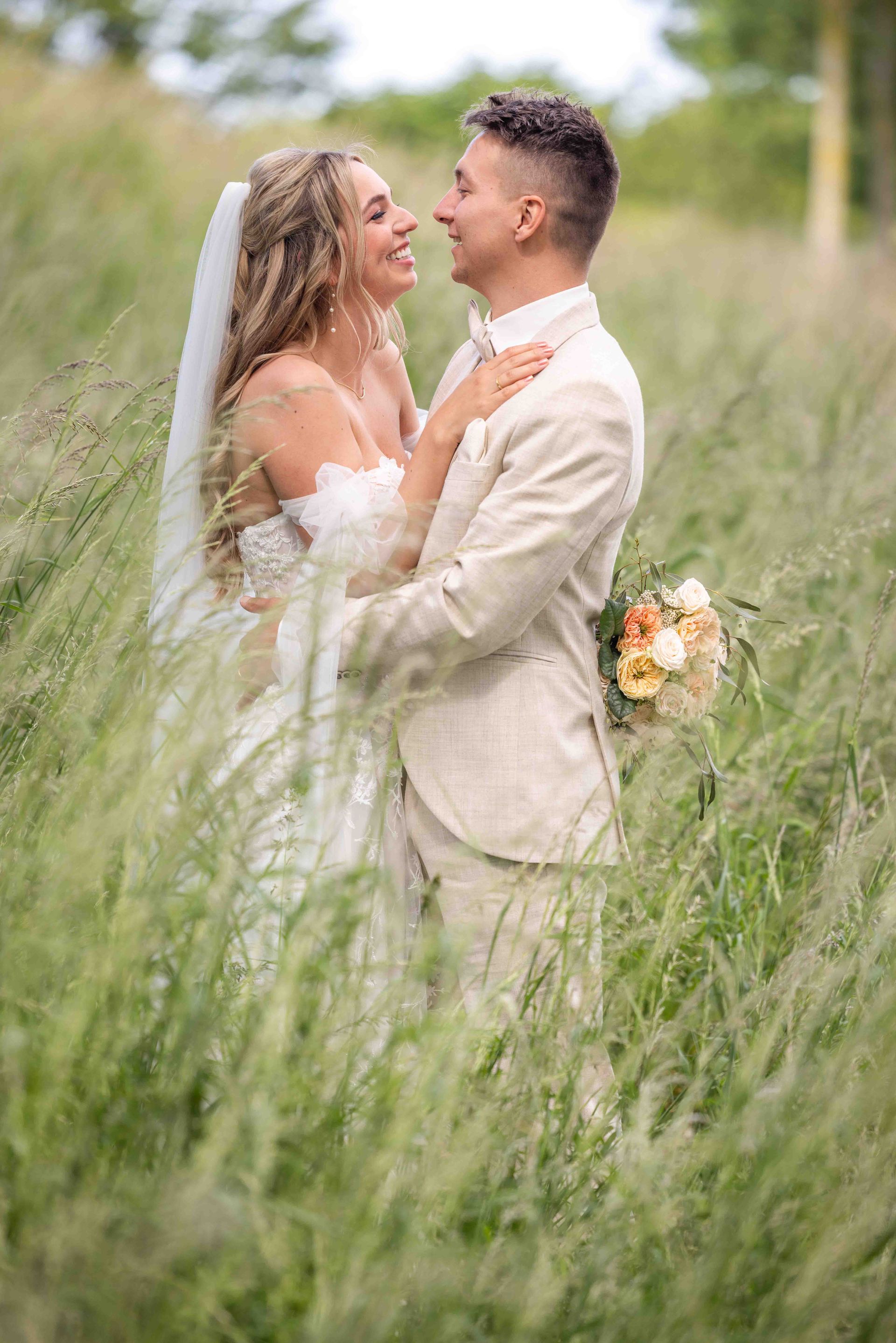Bruid en bruidegom omhelzen elkaar in een veld met hoog gras en glimlachen naar elkaar. De bruid draagt ​​een witte jurk en sluier, de bruidegom een ​​licht pak.