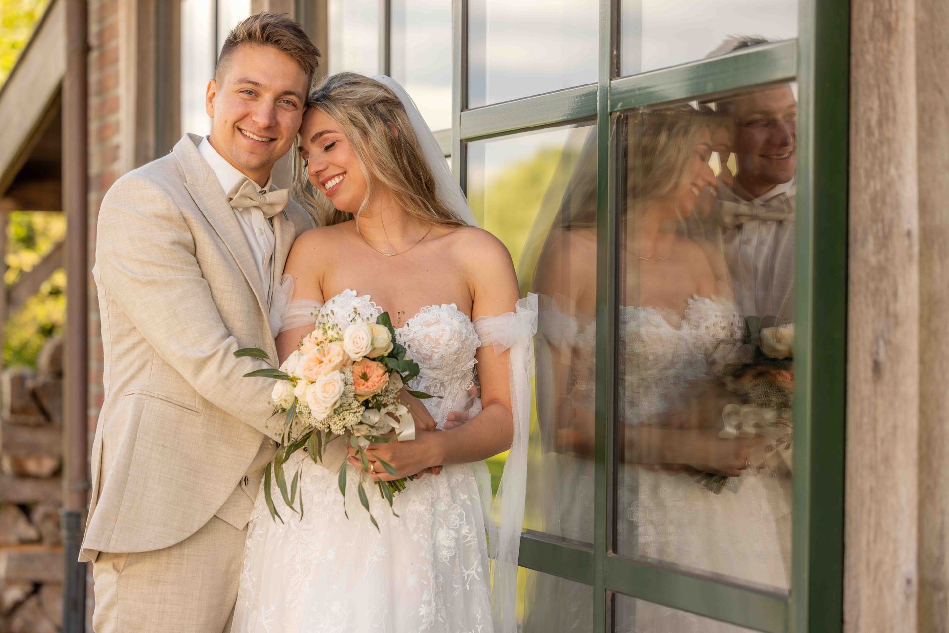 Bruid en bruidegom glimlachen en poseren bij een raam. De bruid draagt ​​een witte jurk, sluier en houdt bloemen vast. De bruidegom draagt ​​een bruin pak.