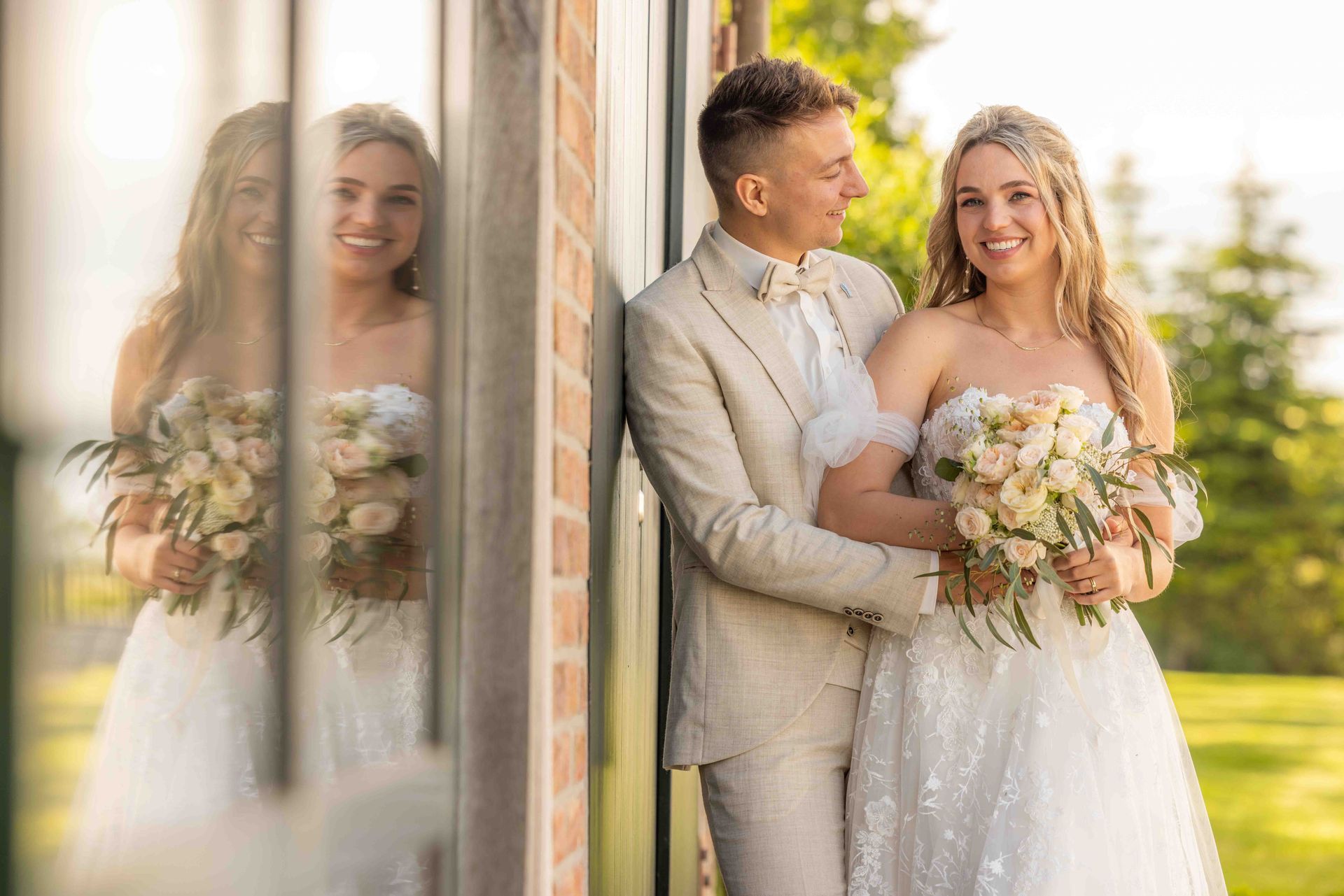 Bruid en bruidegom omhelzen elkaar buiten, met boeketten in de hand. De bruid glimlacht in een witte jurk; de bruidegom in een bruin pak. Weerspiegeling zichtbaar.