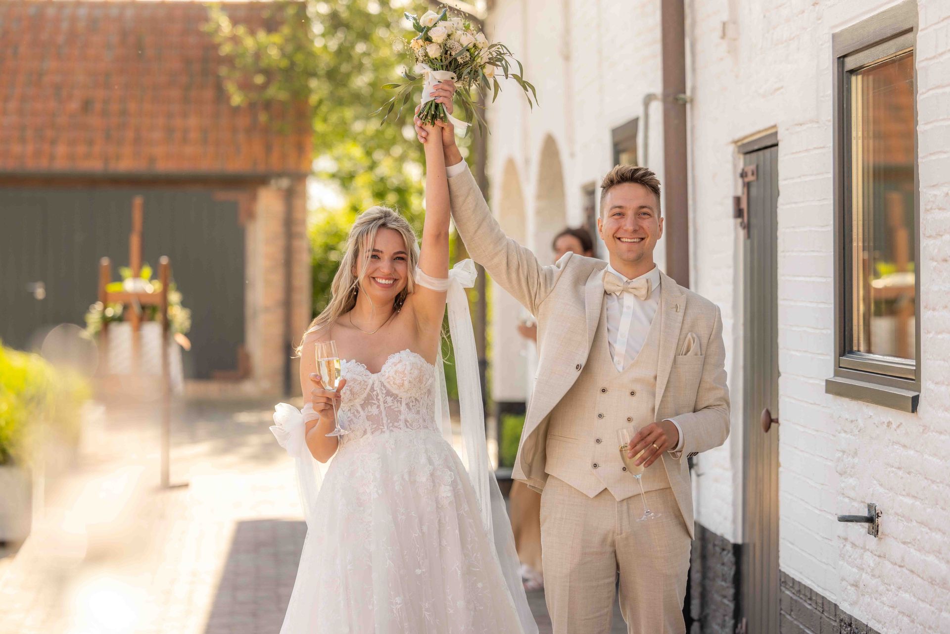 Het bruidspaar viert buiten feest, hand in hand. De bruid draagt ​​een witte jurk, de bruidegom een ​​beige pak.