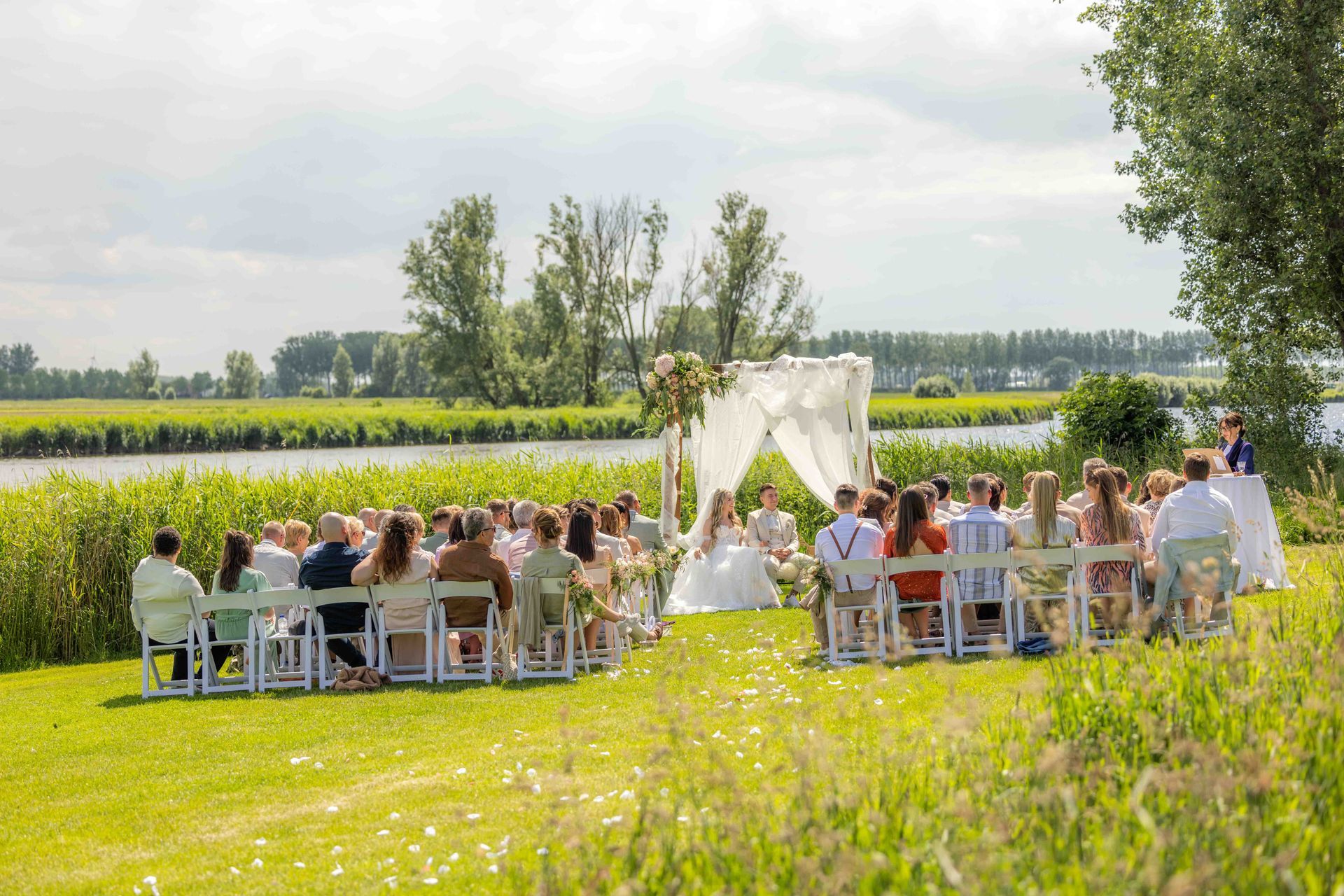 Huwelijksceremonie in de buitenlucht, gasten zitten, bruid en bruidegom bij het altaar. Groen veld, rivier, bewolkte lucht.
