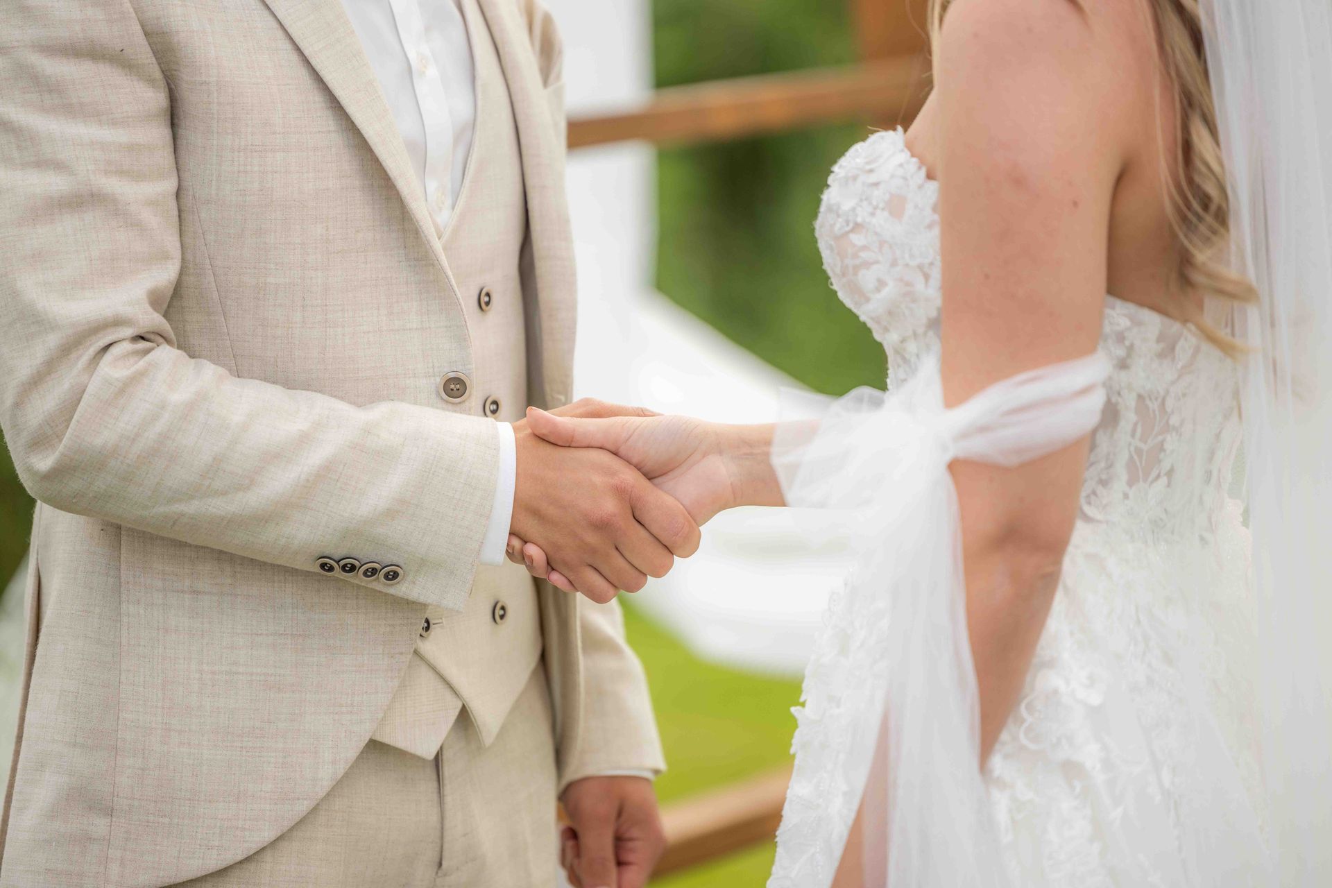 Een stel houdt elkaars hand vast tijdens de huwelijksceremonie. De man draagt ​​een bruin pak, de vrouw draagt ​​een witte jurk.