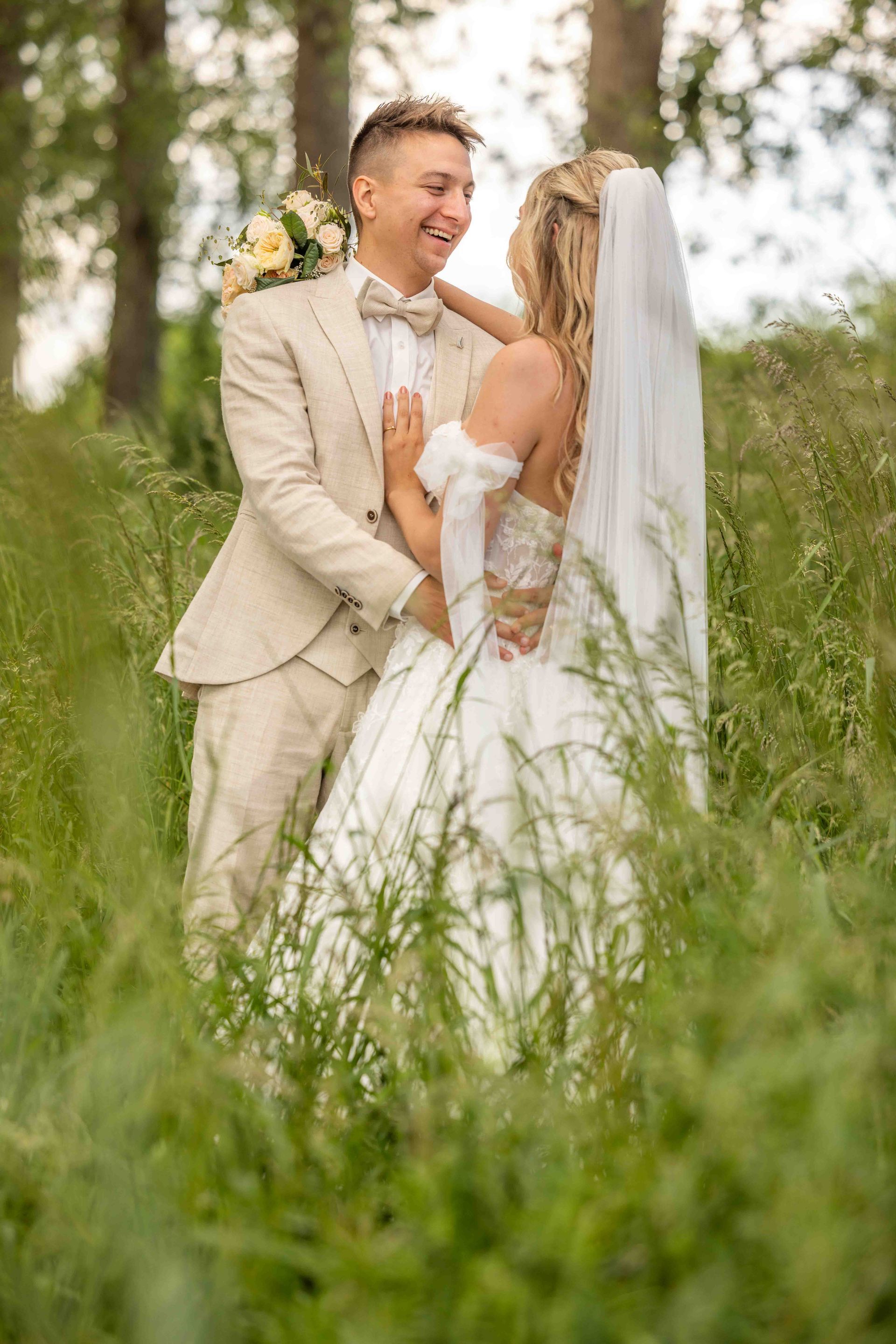 Bruidspaar omhelst elkaar in een veld. De bruidegom in een bruin pak glimlacht naar de bruid in een witte jurk, sluier en met een boeket in haar handen.