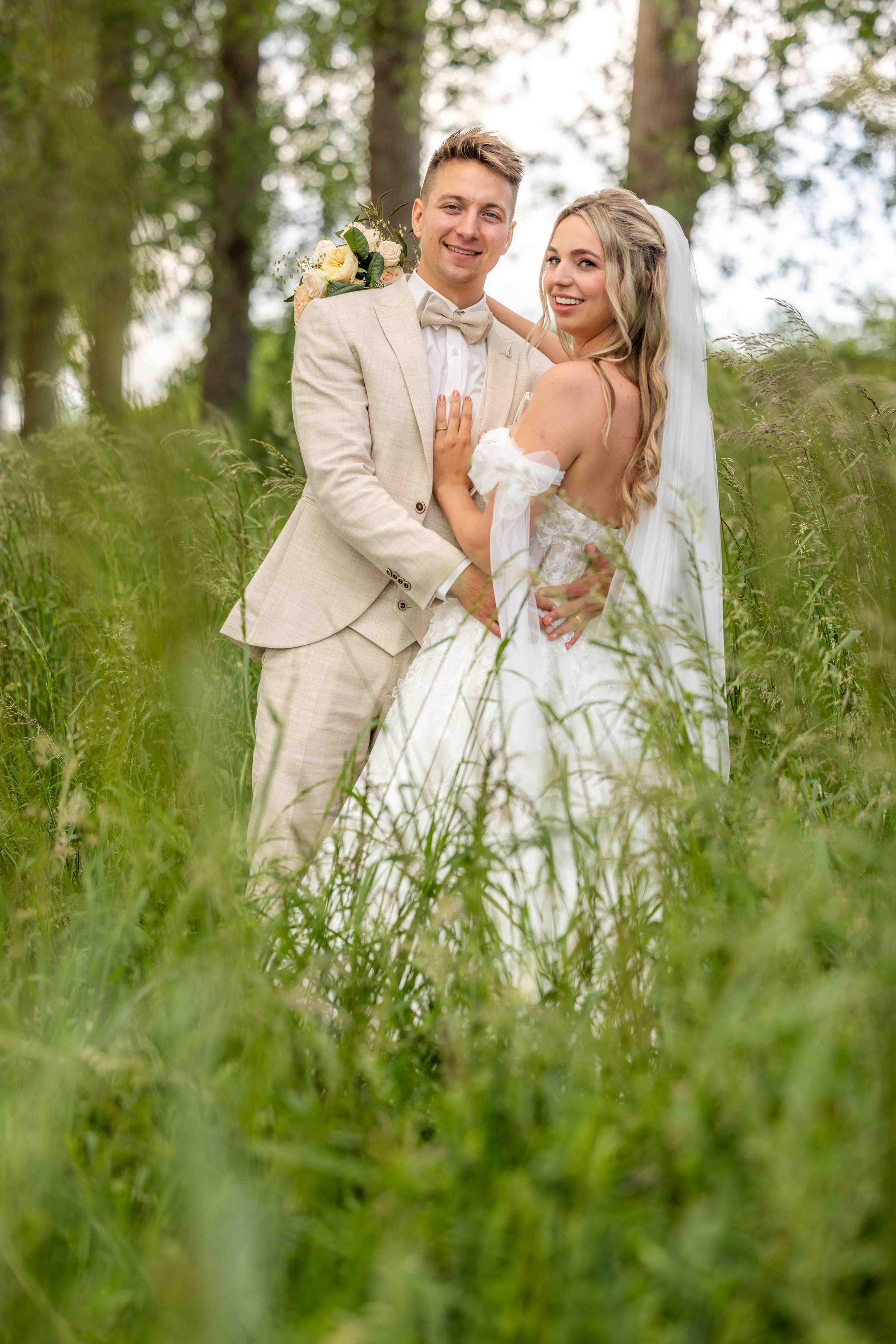 Bruid en bruidegom in formele kleding omhelzen elkaar in een veld met hoog gras. De bruidegom draagt ​​een bruin pak, de bruid een witte jurk.
