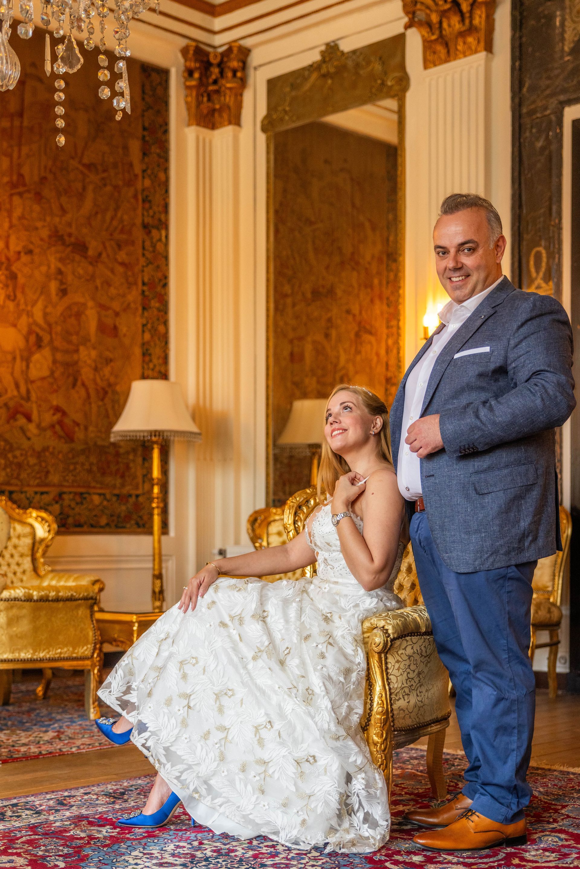 Een vrouw in een jurk, zittend op een sierlijke gouden stoel, kijkt op; een man in een blauw jasje glimlacht en staat naast haar in de vergulde kamer.