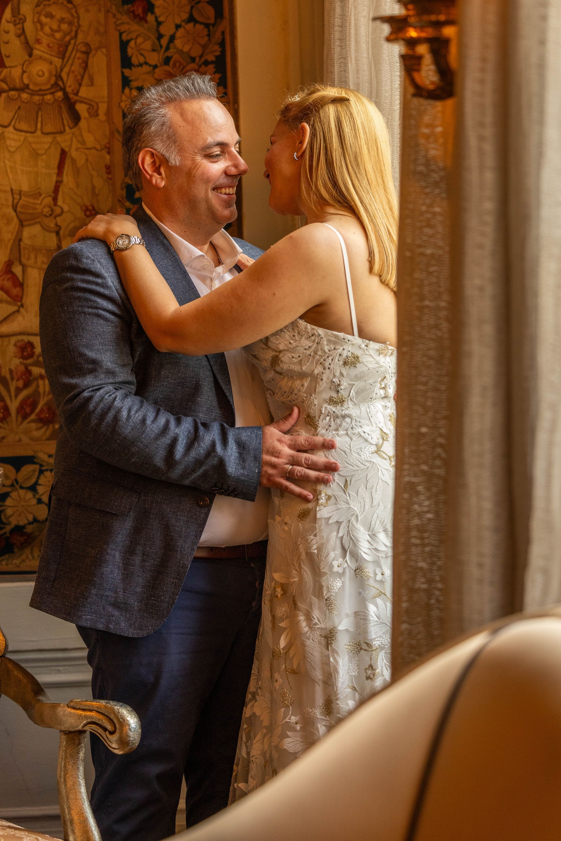 Een paartje omhelst elkaar bij een raam. De vrouw draagt ​​een witte kralenjurk; de man een blazer en een spijkerbroek. Warm licht.