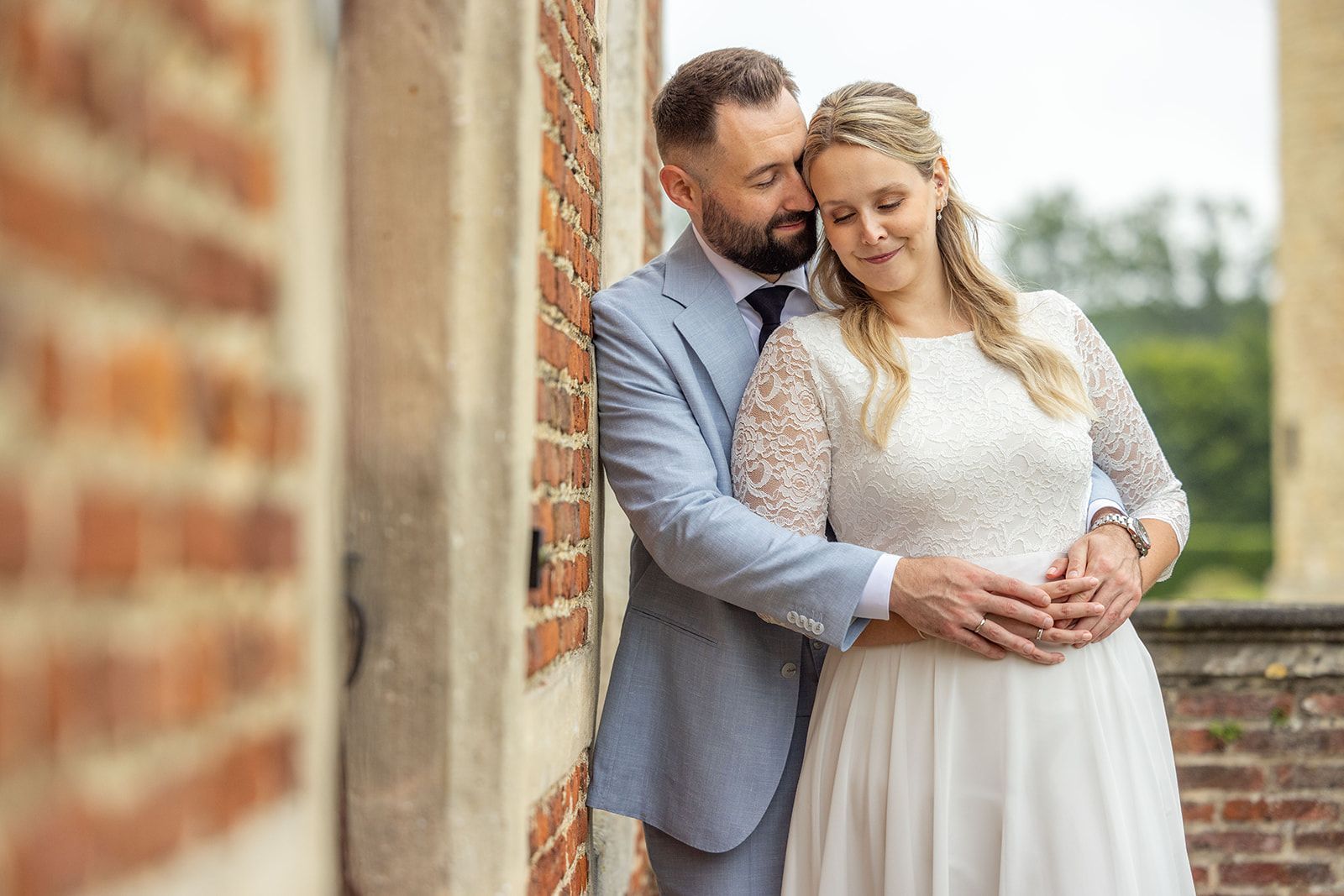 Pasgetrouwden omhelzen elkaar naast een klassieke zwarte auto. De bruid houdt bloemen vast, de bruidegom draagt ​​een rood pak; zonlicht in een voorstedelijke omgeving.