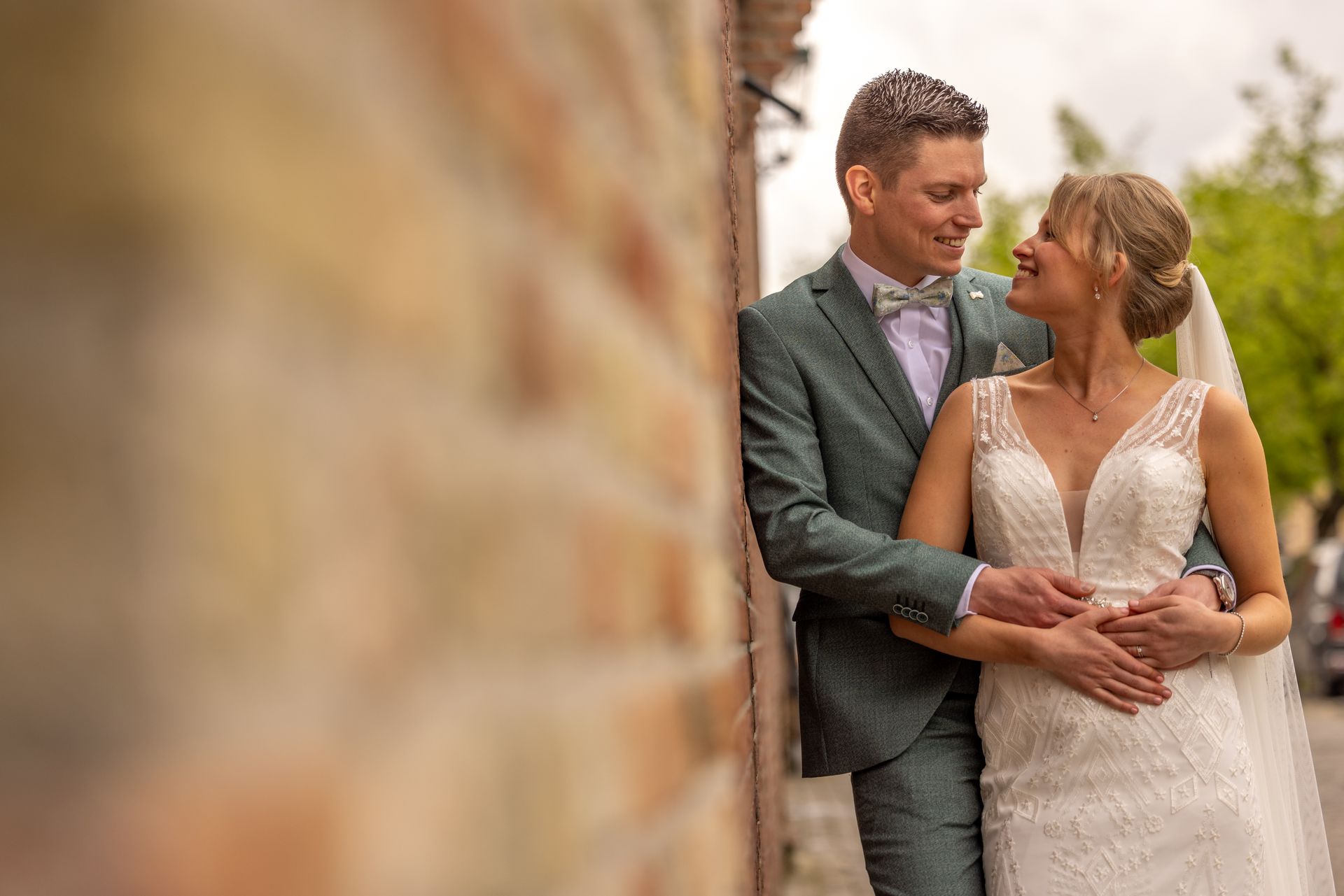Het pasgetrouwde stel omhelst elkaar lachend tegen een bakstenen muur. De bruidegom draagt ​​een groen pak, de bruid een witte jurk.