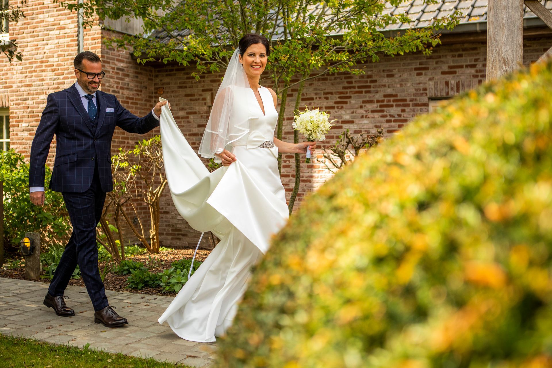 Man in pak helpt bruid met haar jurk terwijl ze naar een bakstenen gebouw lopen, met groen op de voorgrond.