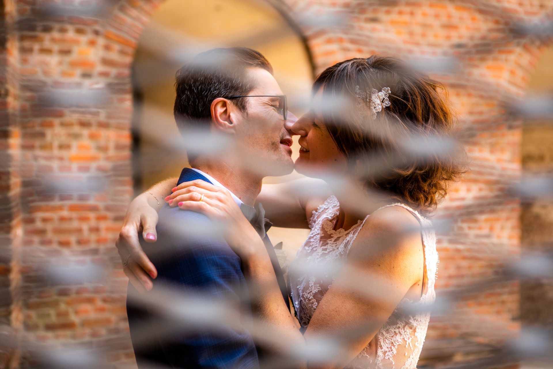 Een paar omhelst elkaar en staat op het punt elkaar te kussen achter een metalen hek, tegen een bakstenen boog.