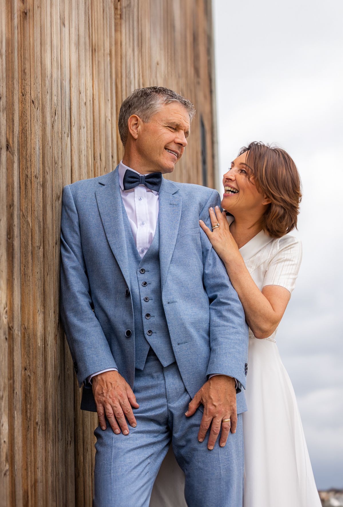 Een liefdevol lachend stel, man in een blauw pak, vrouw in een witte jurk, tegen een houten wand.