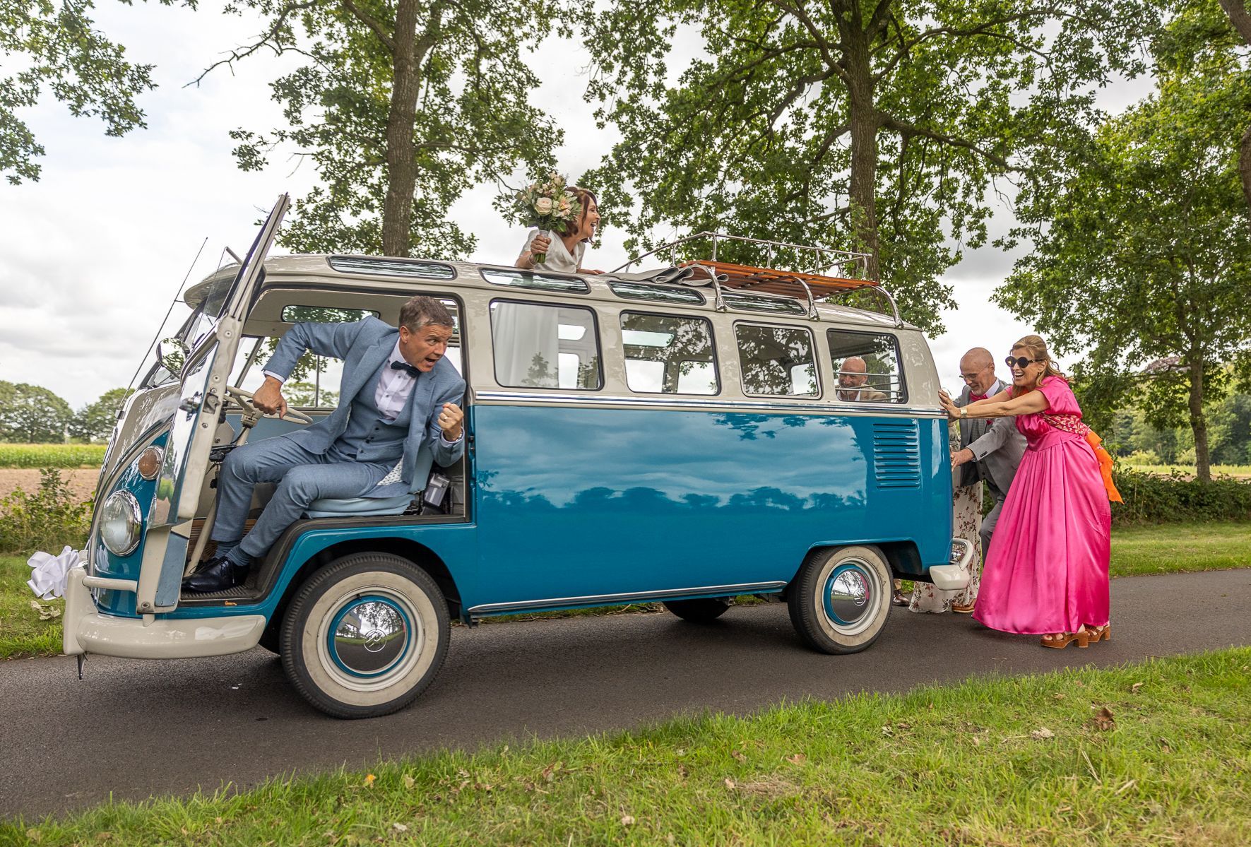 Man in blauw pak probeert uit een blauwgroen VW-busje te komen, terwijl mensen hem van de zijkant duwen, op een weg, tussen het groen.