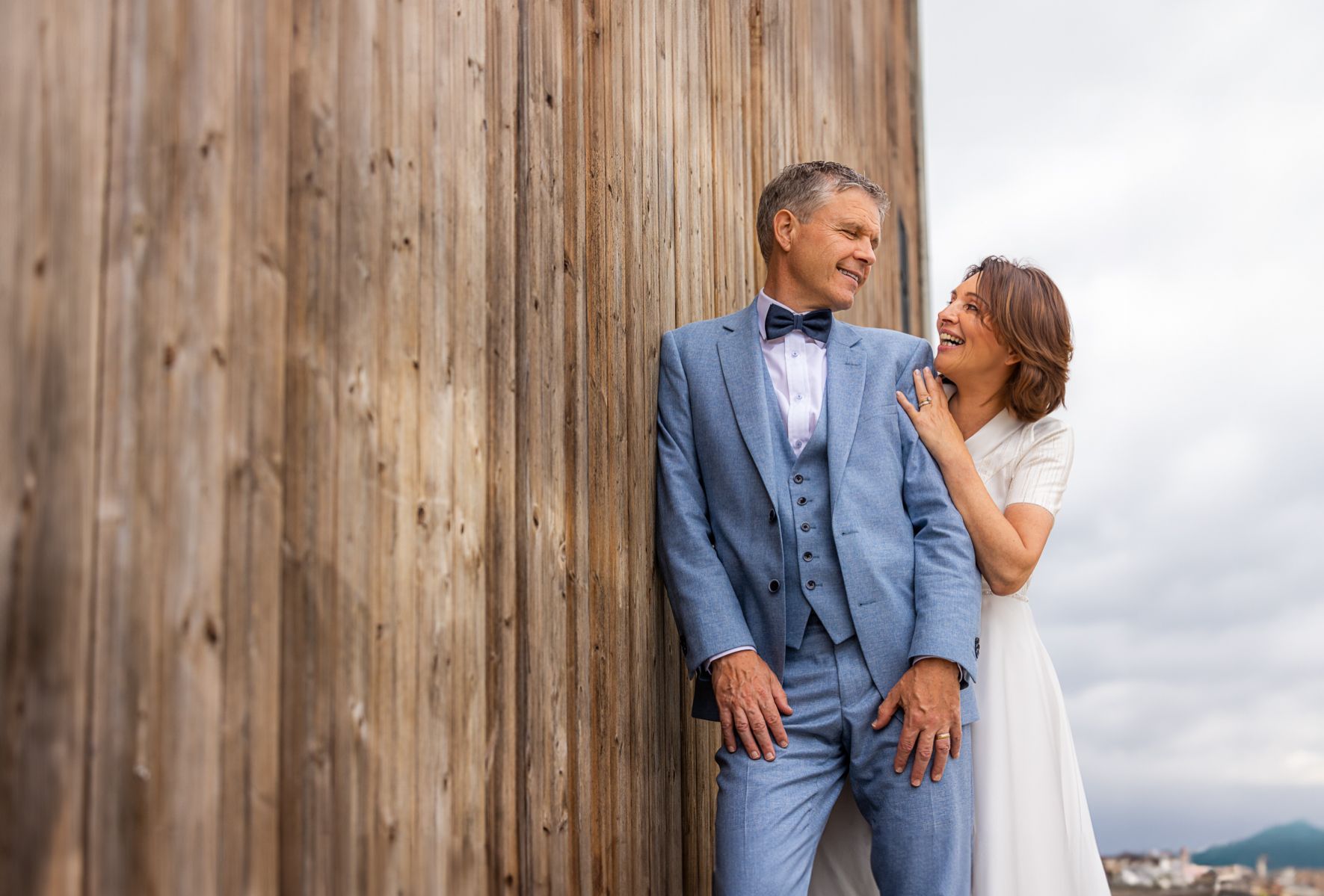 Man in blauw pak en vrouw in witte jurk lachend naast houten lambrisering.