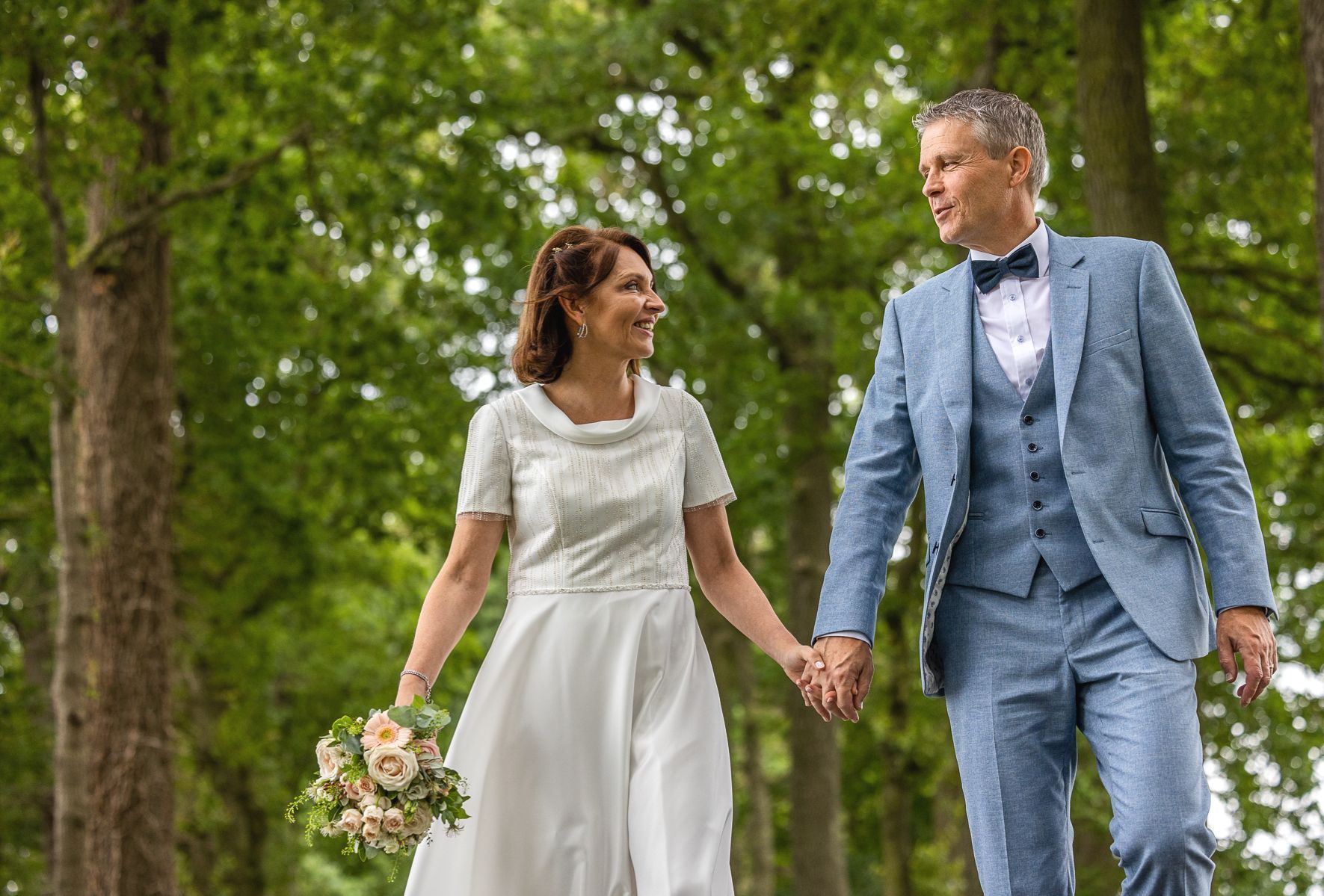 Een stel hand in hand, wandelend in een park, lachend. Bruid in een witte jurk, bruidegom in een blauw pak. Omringd door bomen.