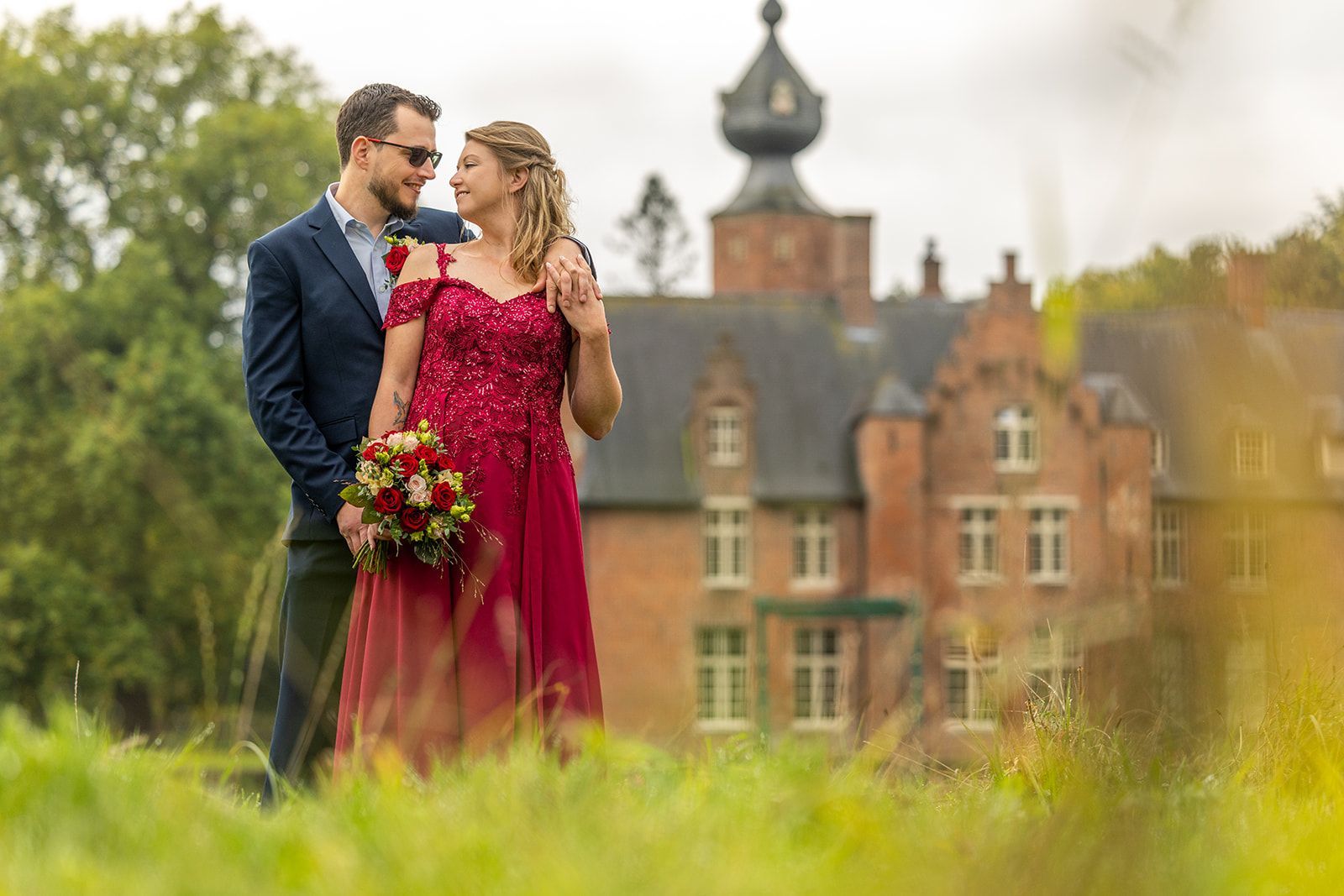 Couple in wedding attire embrace in front of a castle, bride in red dress, groom in blue suit.