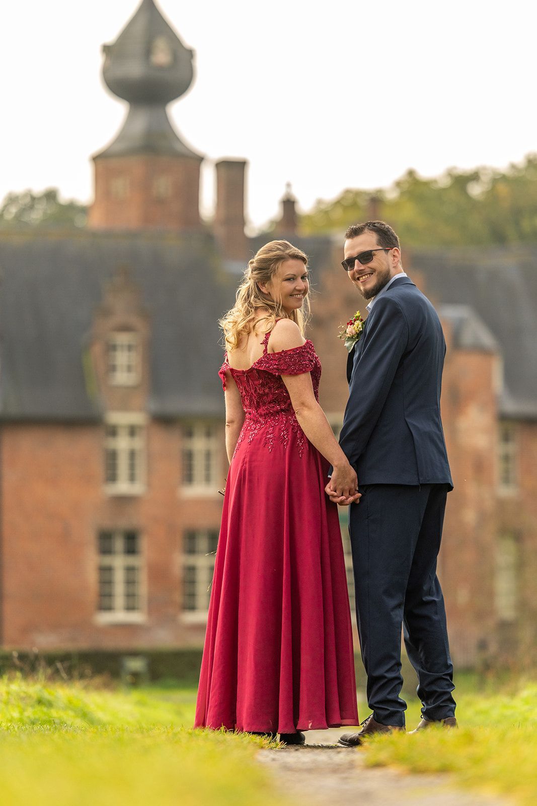 Een echtpaar in formele kleding houdt elkaars hand vast en kijkt over hun schouders naar de camera voor een bakstenen gebouw.