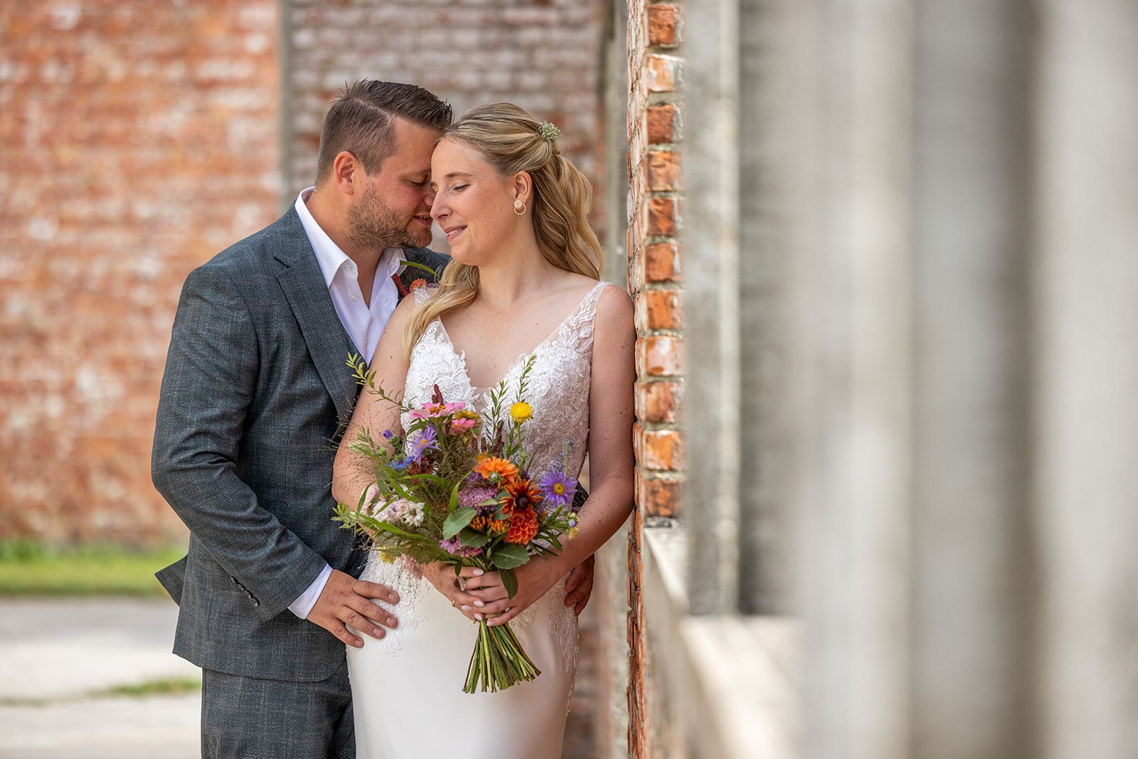 Bruid en bruidegom omhelzen elkaar, houden bloemen vast en leunen tegen een bakstenen muur. Het is een zonnig buitentafereel.