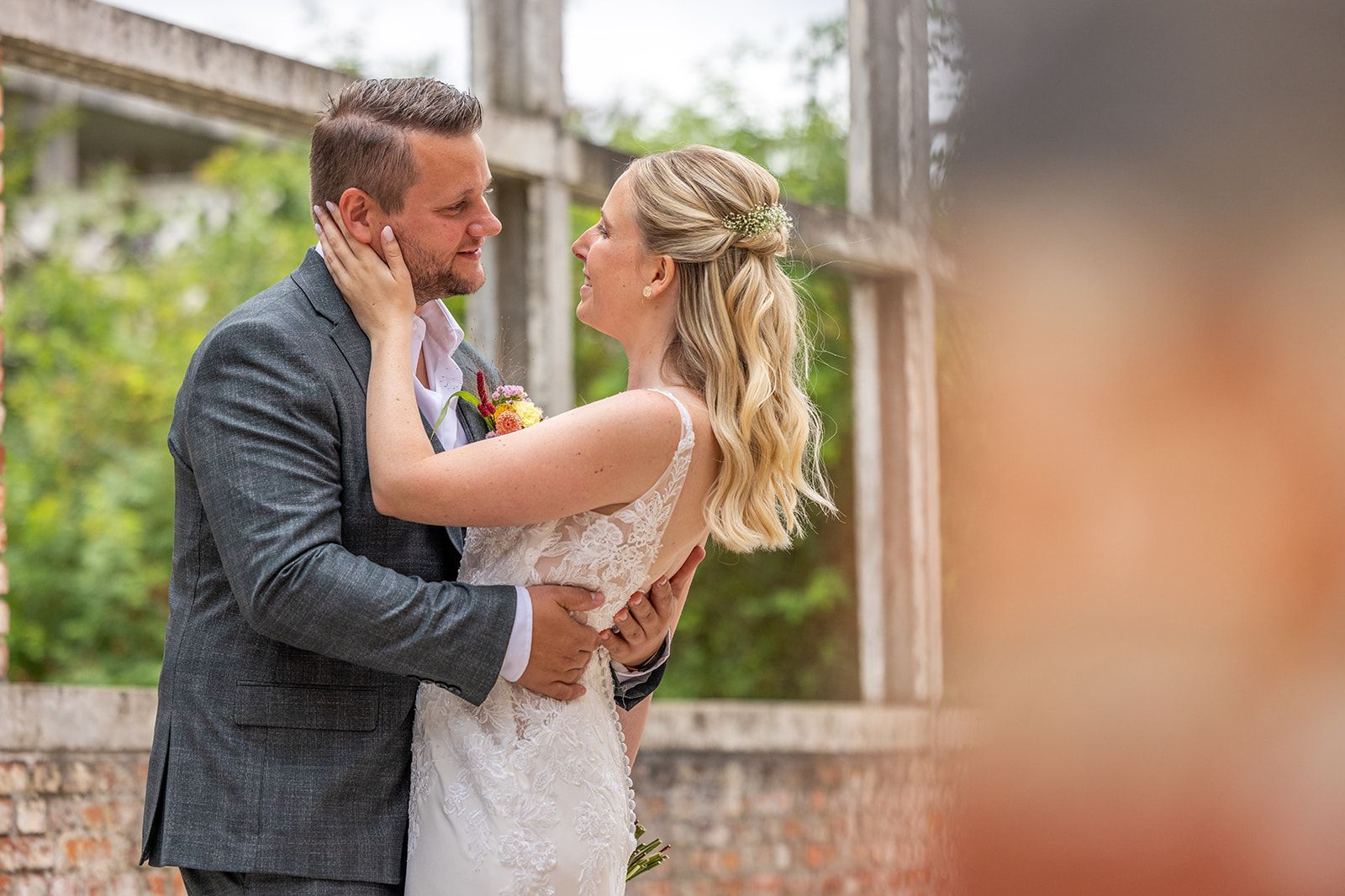Het paar omhelst elkaar lachend voor een gebouw van baksteen en beton. De bruid draagt ​​een witte jurk. De bruidegom een ​​grijs pak.