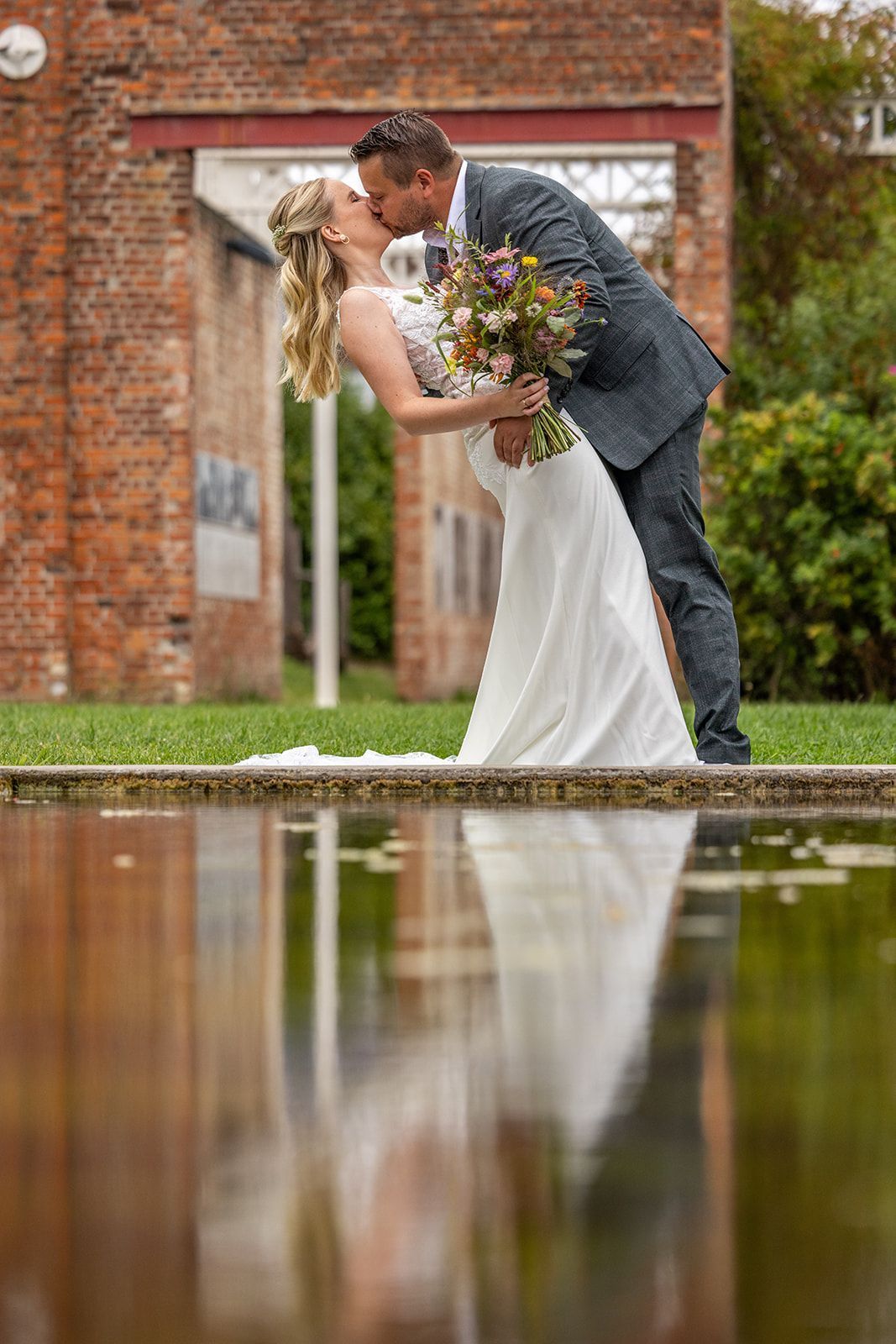 Bruid en bruidegom kussen elkaar bij een spiegelend zwembad, met een boeket in hun handen, tegen een bakstenen gebouw.