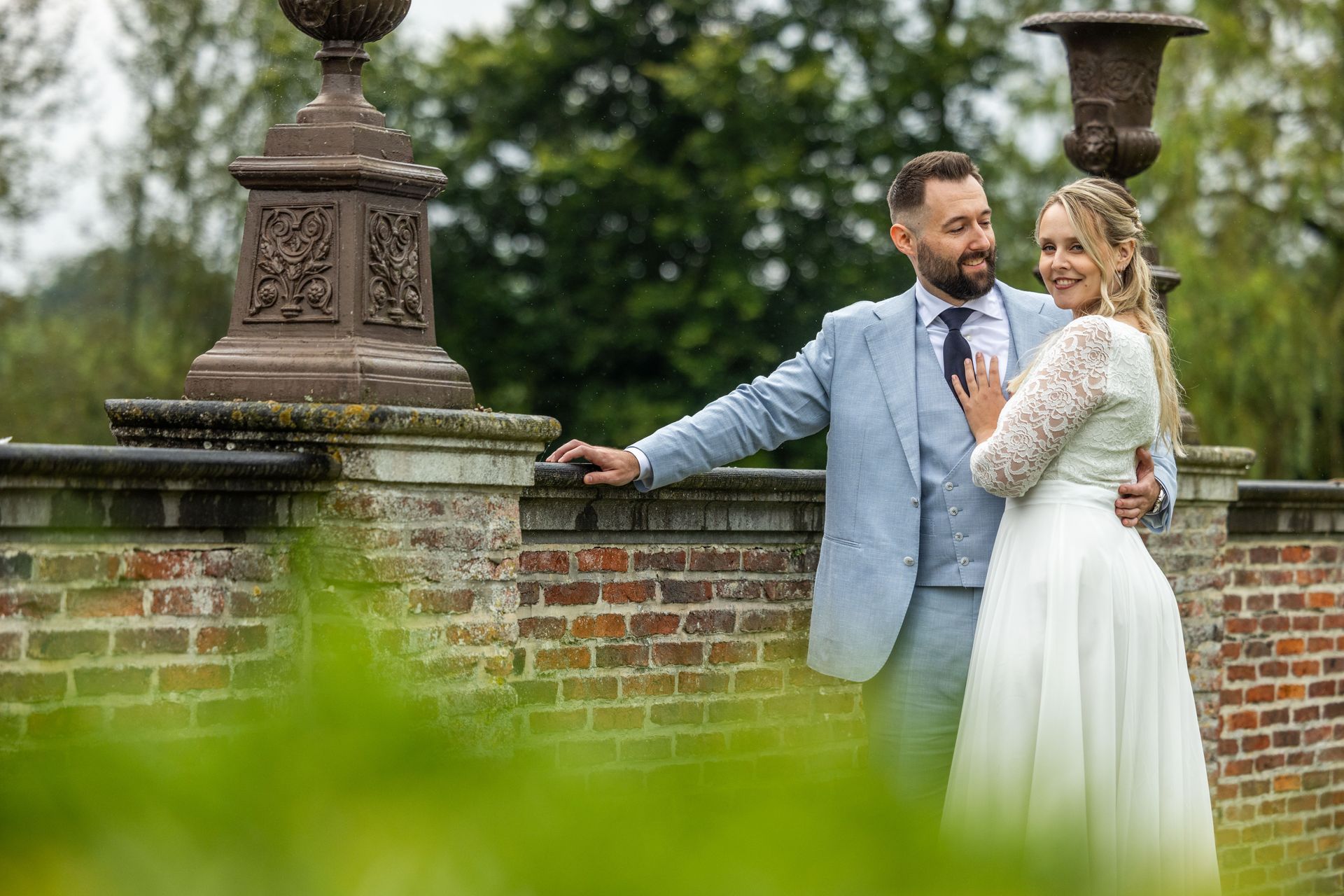 Het pasgetrouwde stel poseert bij een bakstenen muur en sierlijke lantaarnpalen; de bruid glimlacht terwijl de bruidegom haar omhelst.