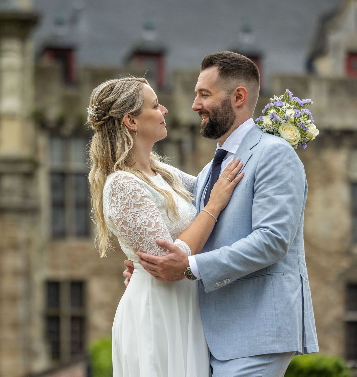 Bruid en bruidegom omhelzen elkaar en kijken elkaar aan, buiten. Zij draagt ​​een witte jurk; hij een blauw pak.