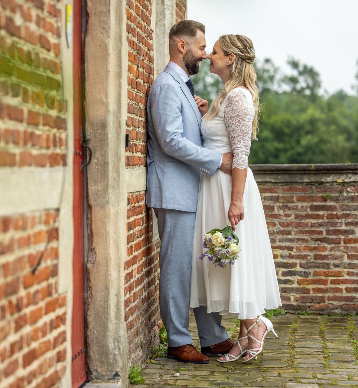 Bruidspaar, man in blauw pak en vrouw in witte jurk, leunen tegen een bakstenen muur en staan ​​op het punt om te kussen.