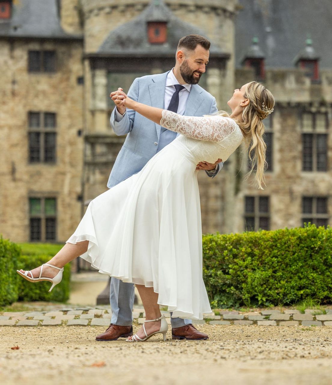 Dansend paar buiten, bruid in witte jurk, bruidegom in blauw pak, historisch kasteel op de achtergrond.