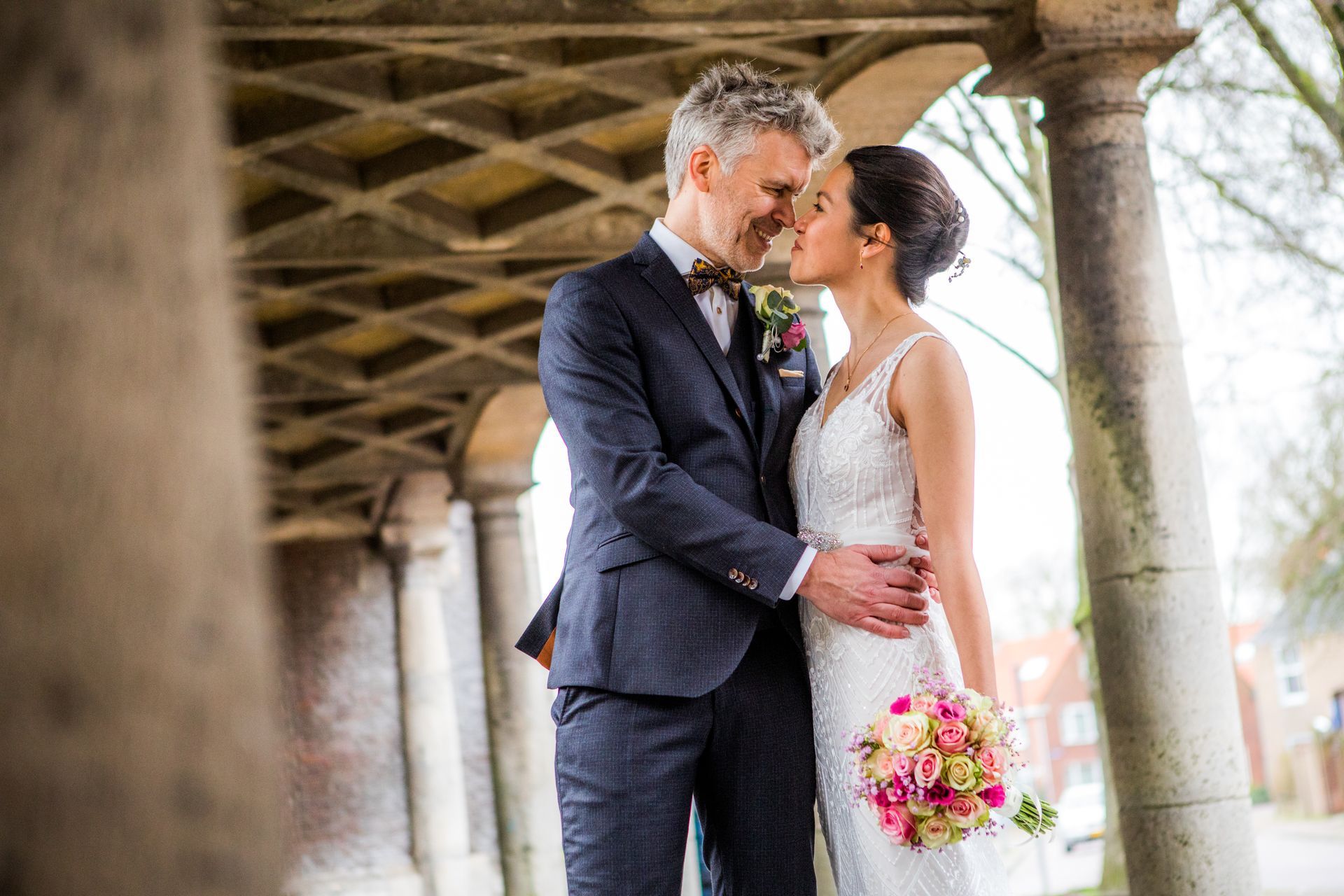 Bruid en bruidegom omhelzen elkaar onder een stenen arcade, lachend, boeket.