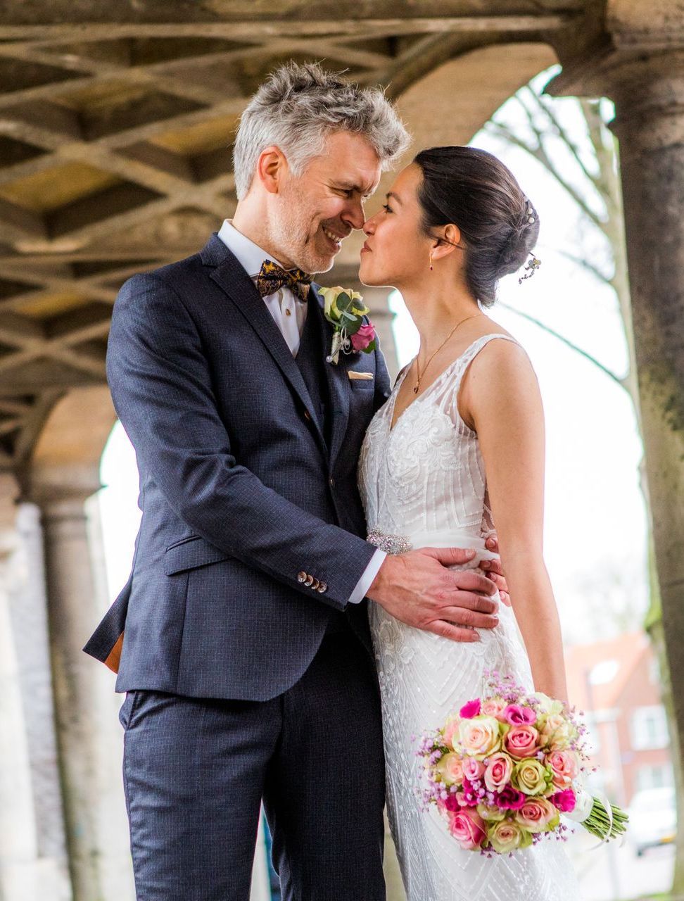 Een paar omhelst elkaar op een bruiloft. De man draagt een pak en de vrouw een witte jurk, met een boeket in haar handen, onder een boogvormige loopbrug.