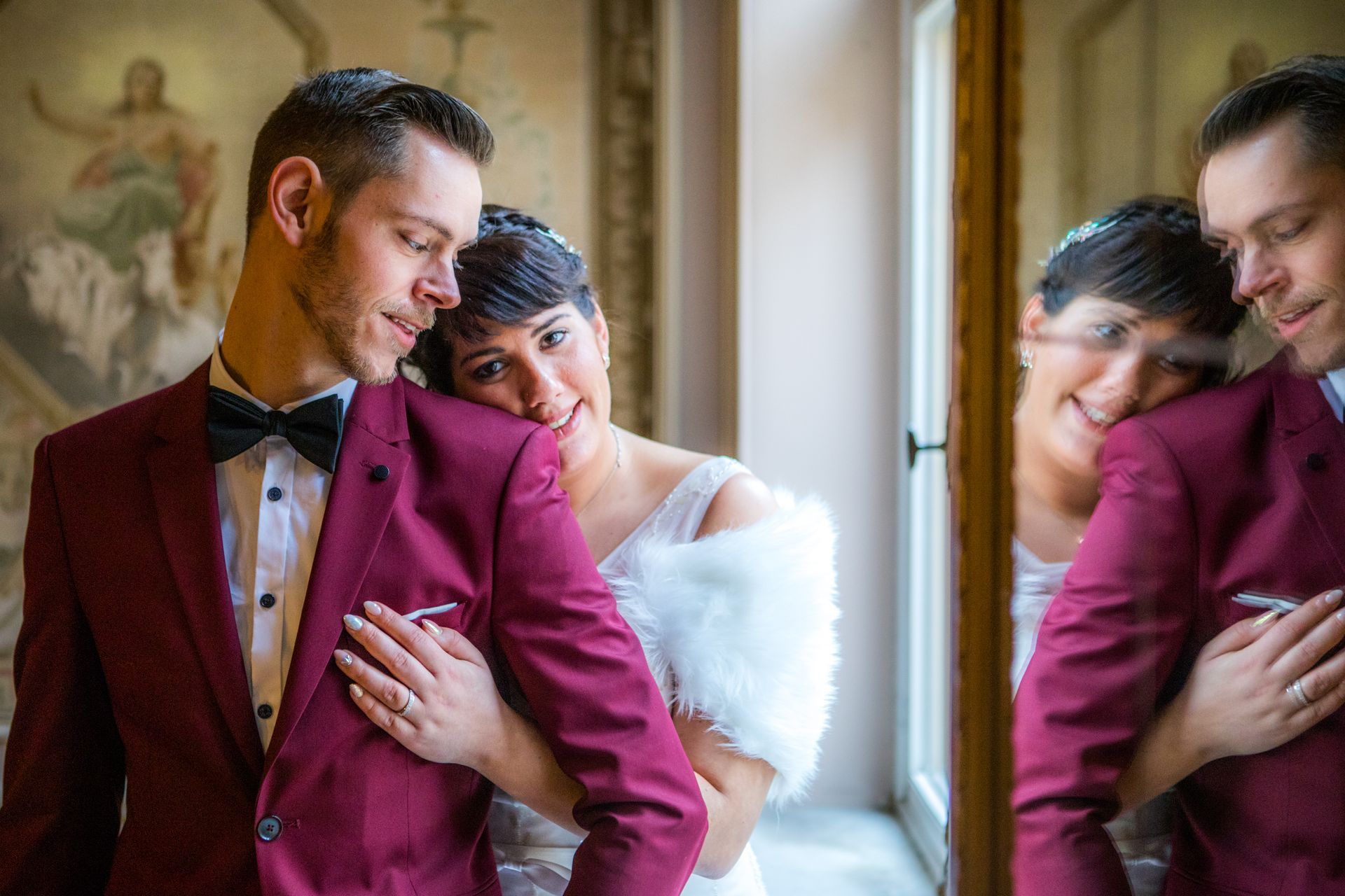 Bruid en bruidegom omhelzen elkaar op de trouwdag. Bordeauxrood pak, witte jurk, bontjas, glimlachen, spiegelbeeld.