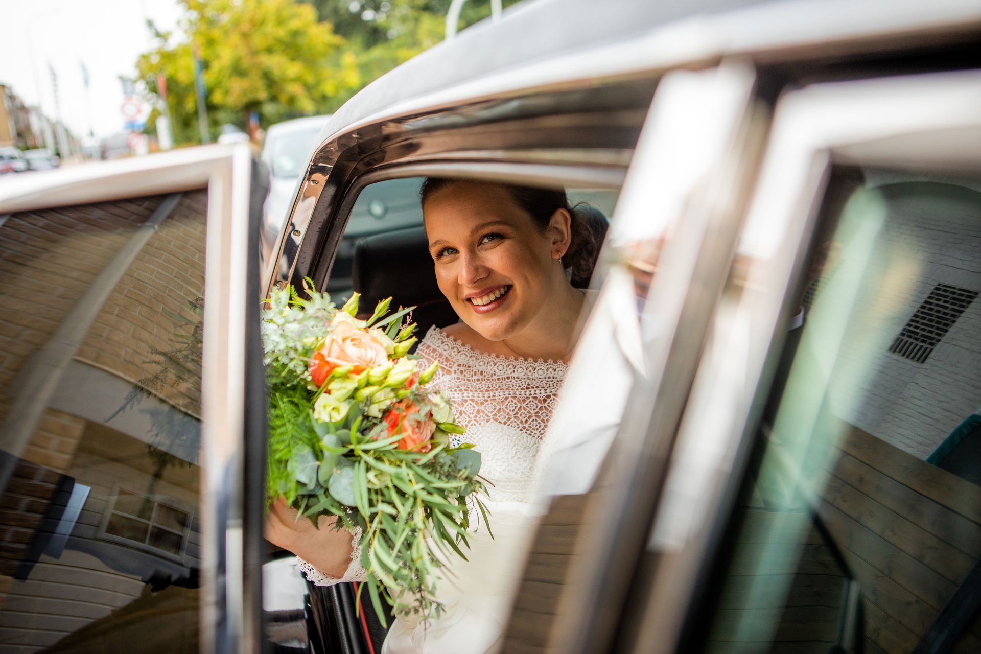 Bruid in een trouwjurk, met bloemen in de hand, glimlachend in de auto met open deuren.