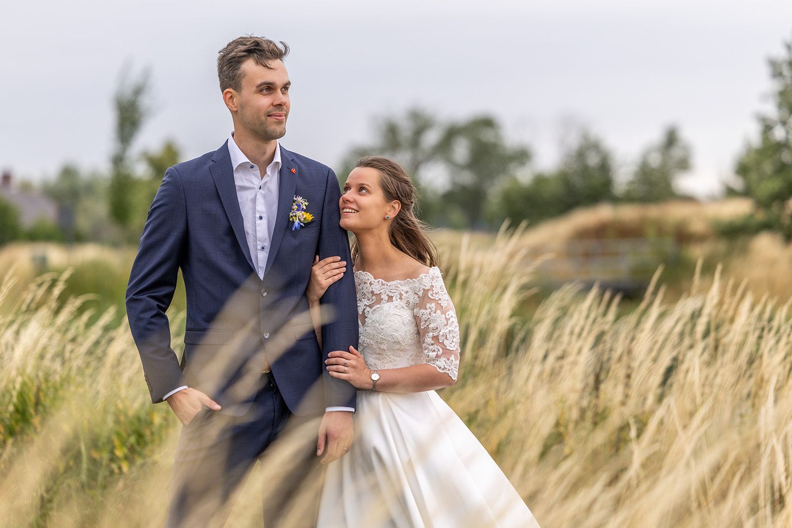 Bride and groom in wedding attire, embracing outdoors amid tall grass. Cloudy sky backdrop.