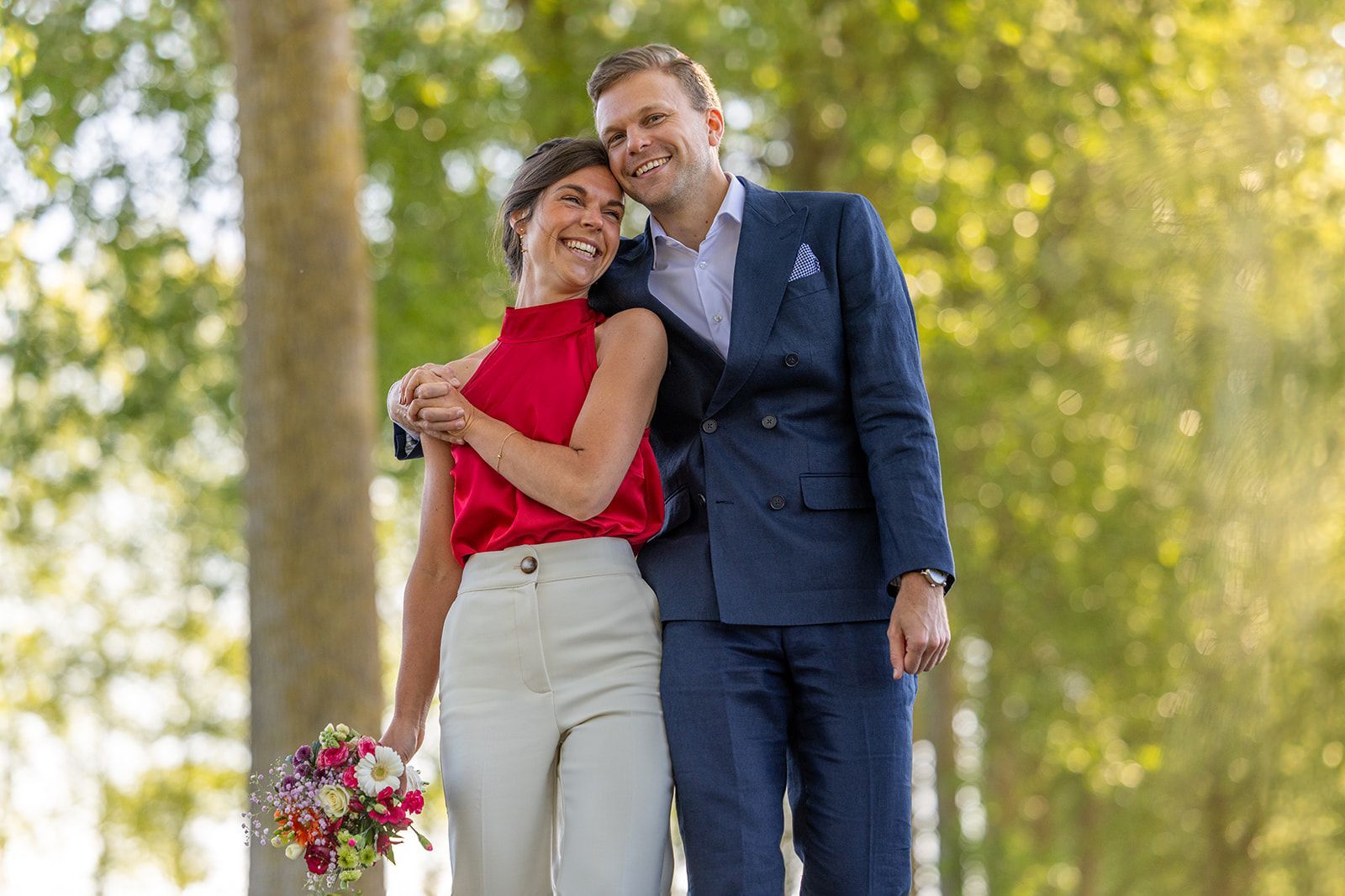 Pasgetrouwden omhelzen elkaar naast een klassieke zwarte auto. De bruid houdt bloemen vast, de bruidegom draagt ​​een rood pak; zonlicht in een voorstedelijke omgeving.