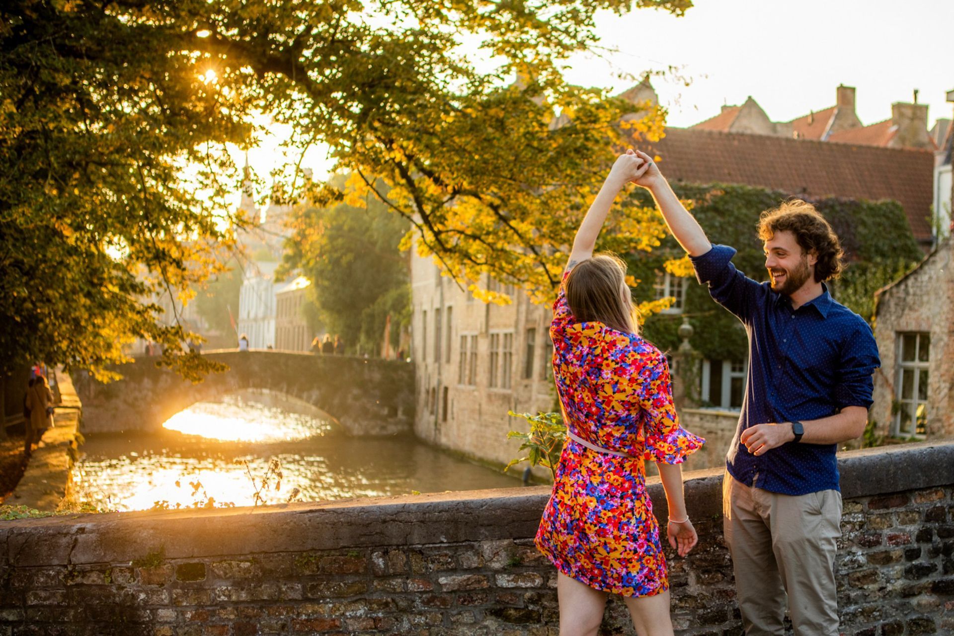 Een stel houdt elkaars hand vast en danst bij een kanaal, met gouden zonlicht in Brugge, België.