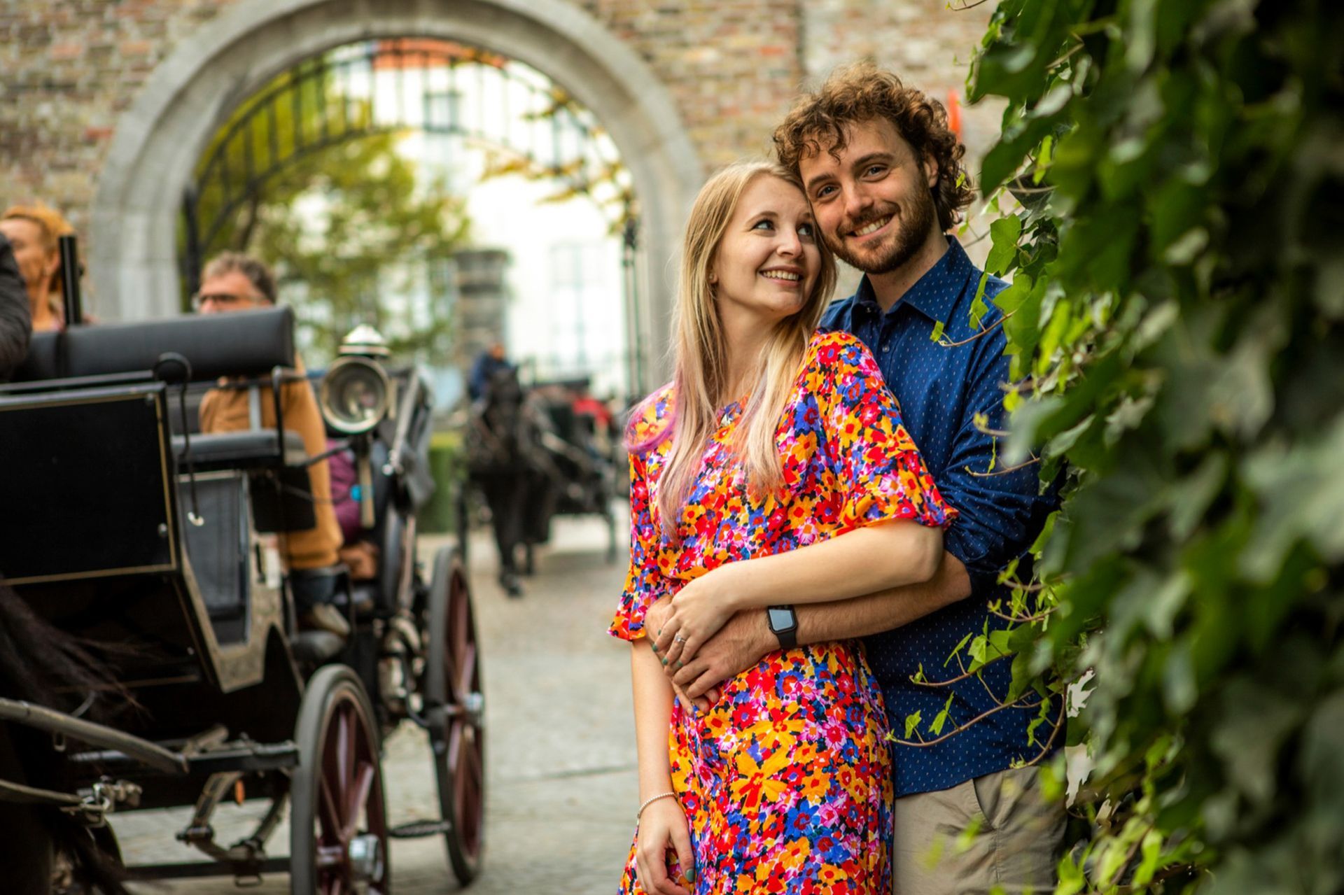 Een paar omhelst elkaar onder een boog en glimlacht naar de camera. Op de voorgrond staat een door paarden getrokken koets.