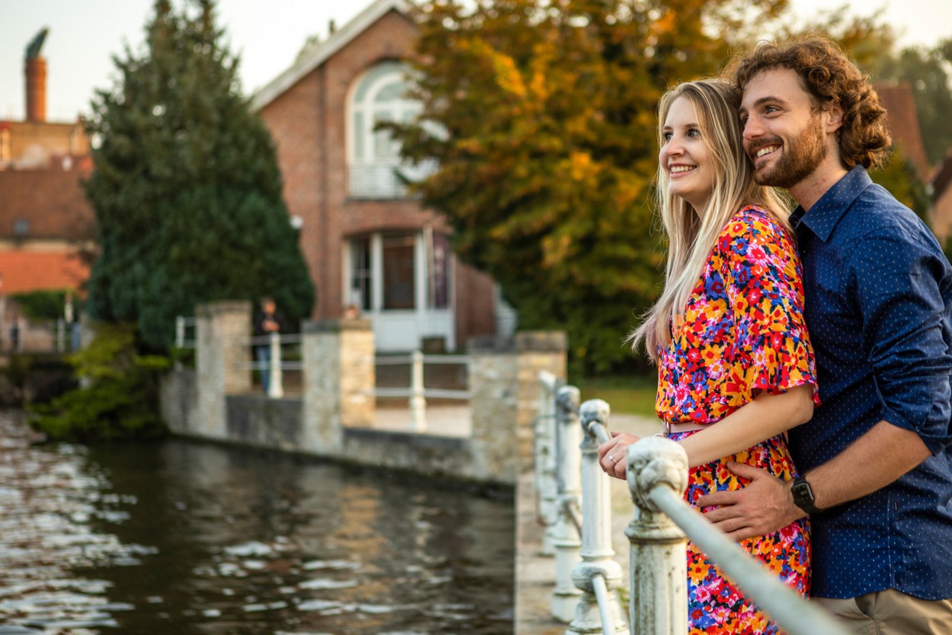 Een lachend stel staat op een brug met uitzicht op een kanaal voor bakstenen gebouwen met bomen.