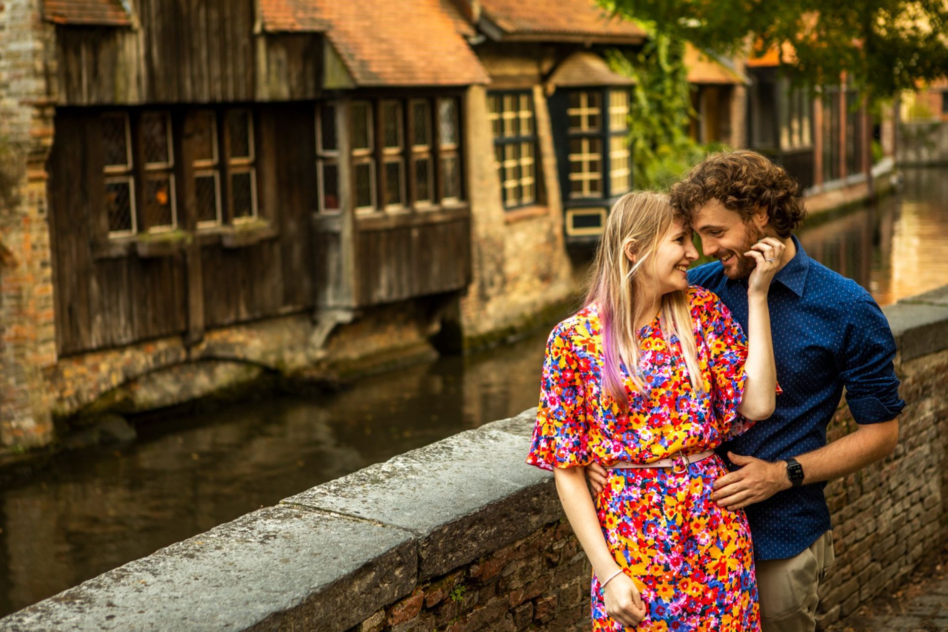 Een paar omhelst elkaar bij een kanaal; de vrouw draagt een bloemenjurk, de man draagt een blauw shirt. Op de achtergrond staan historische gebouwen.