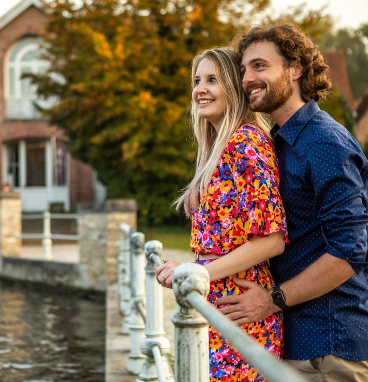 Een lachend stel kijkt uit over het landschap bij een kanaal. De vrouw draagt ​​een bloemenjurk; de man heeft een baard en een blauw shirt.