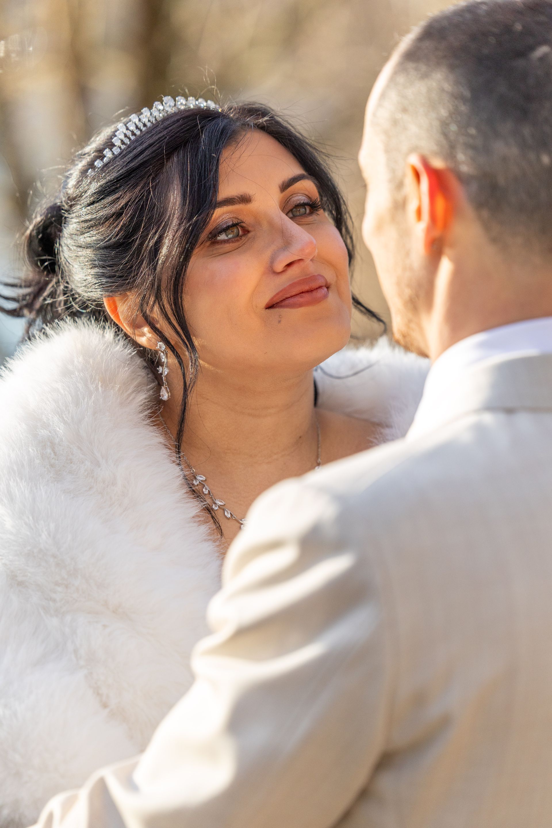 Bride looking at groom, wearing a white fur shawl, outdoors, soft focus, sunny.