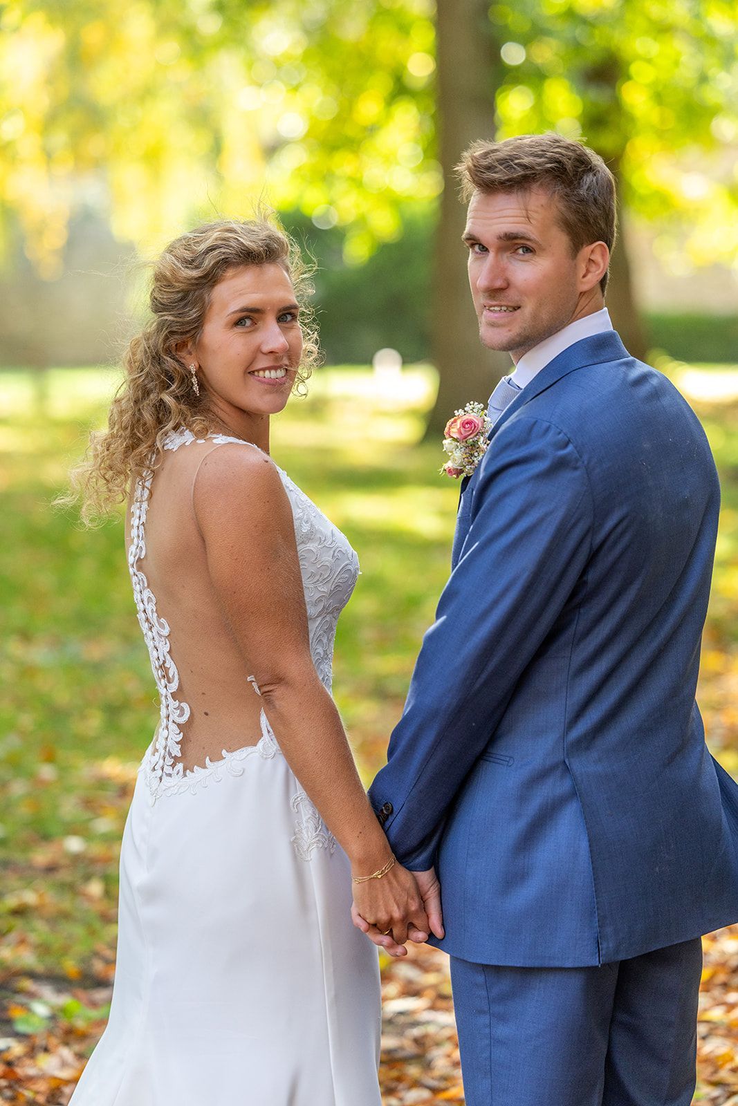 Bruid en bruidegom hand in hand, lachend. Zij draagt een witte jurk, hij een blauw pak, buiten tussen de bomen.
