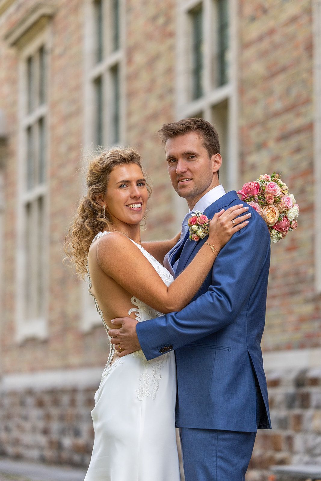 Bruid en bruidegom omhelzen elkaar buiten, met een boeket in de hand. De vrouw draagt een witte jurk; de man een blauw pak.