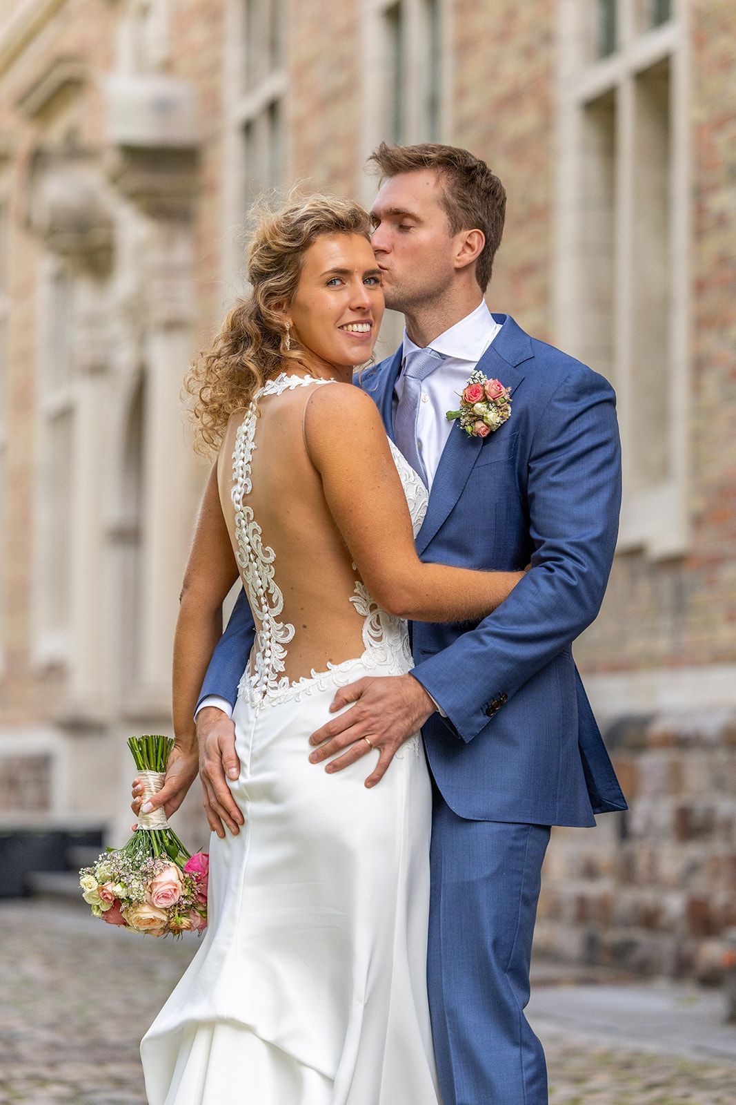 Bruid in witte jurk poseert met bruidegom in blauw pak terwijl hij haar hoofd kust.
