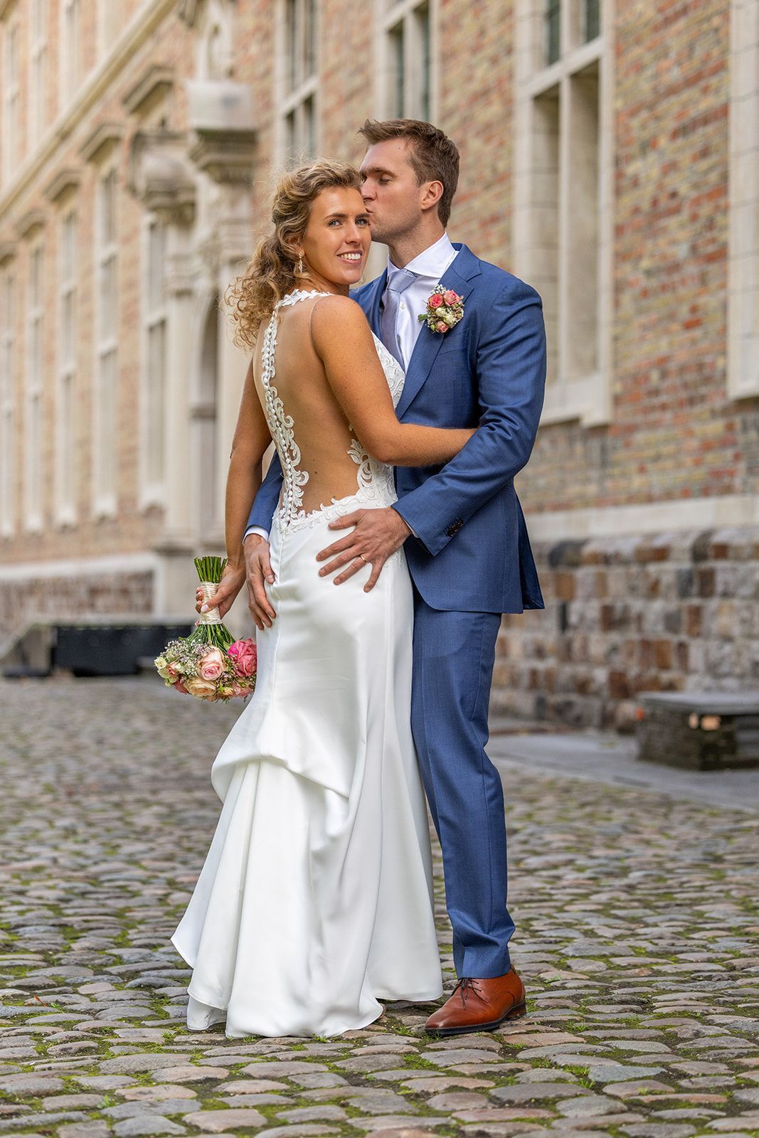 Bruidspaar poseert, hij kust haar voorhoofd. Bruid draagt een witte jurk, bruidegom een blauw pak. Straat met kinderkopjes en een bakstenen gebouw op de achtergrond.