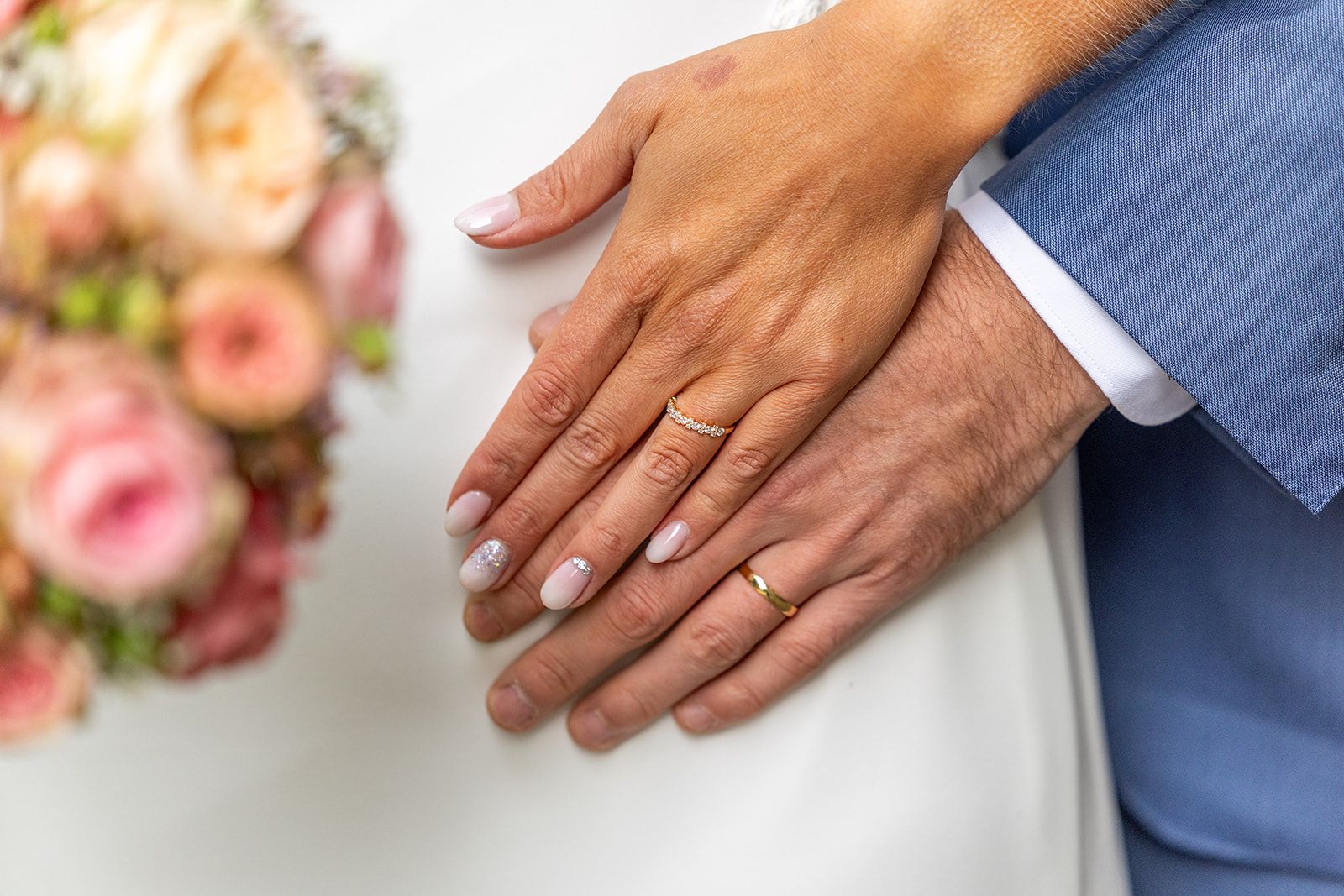 Handen met trouwringen, de een boven de ander, bij een boeket en een blauw pak.