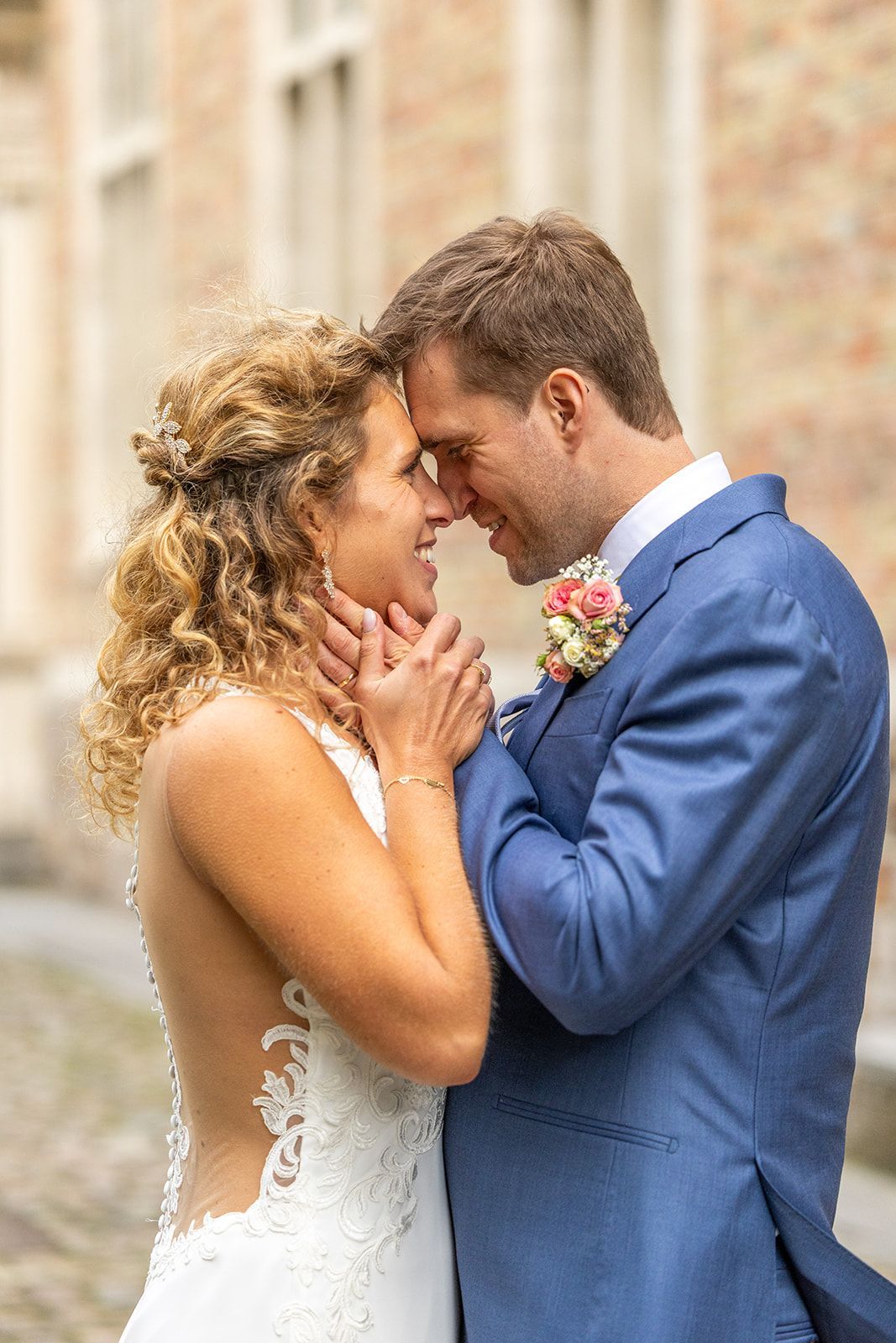 Bruid en bruidegom omhelzen elkaar en raken elkaars voorhoofd aan. De bruid in een witte jurk, de bruidegom in een blauw pak, buiten.